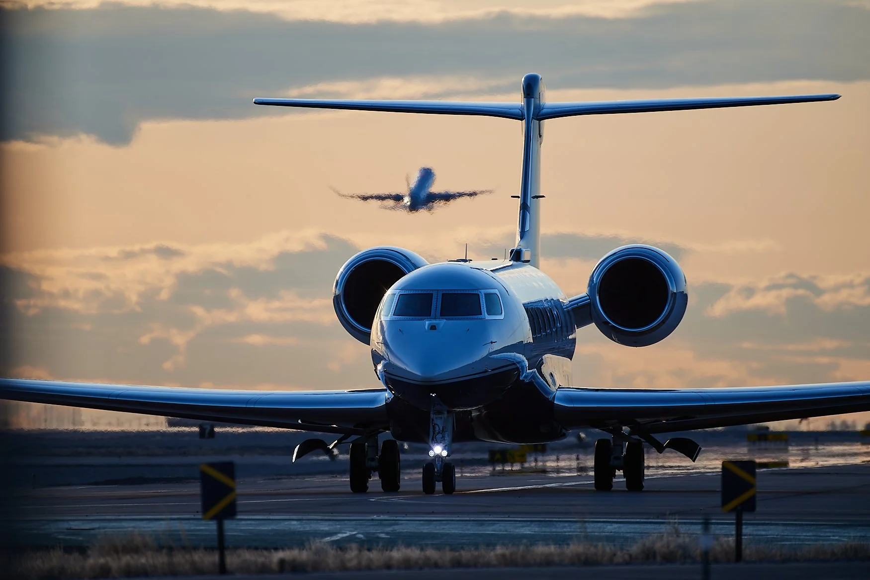 A front view of a private jet on an airport runway with another jet flying in the background during sunset.