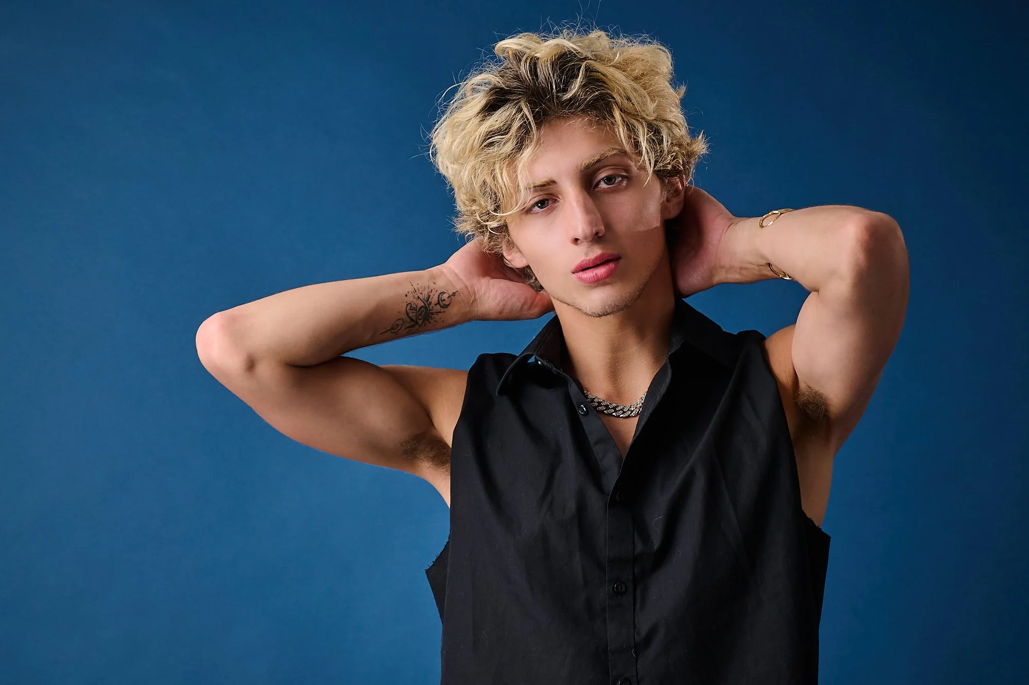 A young person with curly blond hair posing against a blue background, wearing a black sleeveless shirt, gold jewelry, and a tattoo on their left forearm.