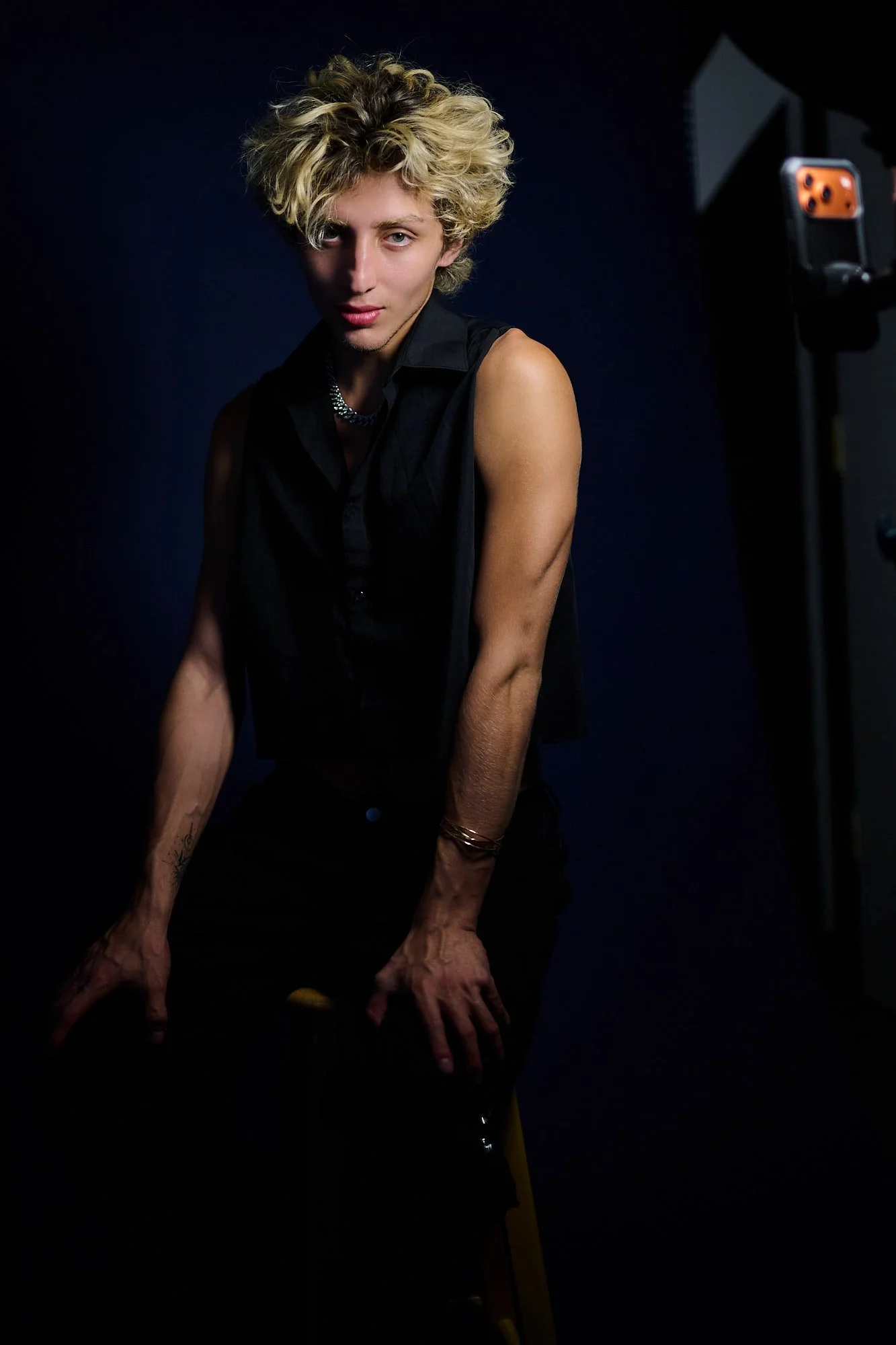 A young man with short, curly blonde hair and wearing a black sleeveless shirt, posing in front of a dark background with professional lighting equipment visible on the right side of the image.