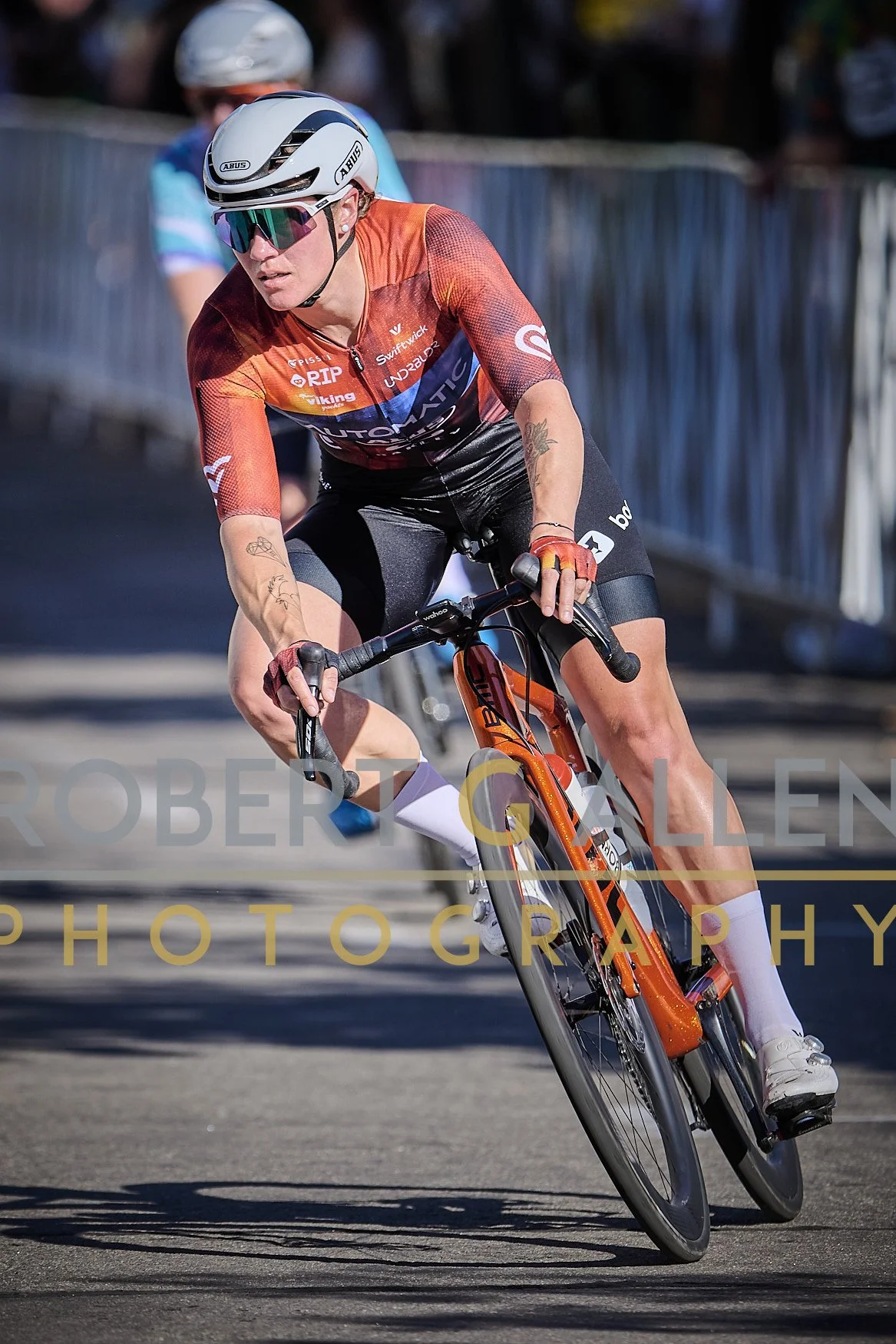 A female cyclist in an orange and black jersey, wearing a white helmet and mirrored sunglasses, leans into a turn on a mountain bike during a race, with a blurred background and another cyclist behind her.