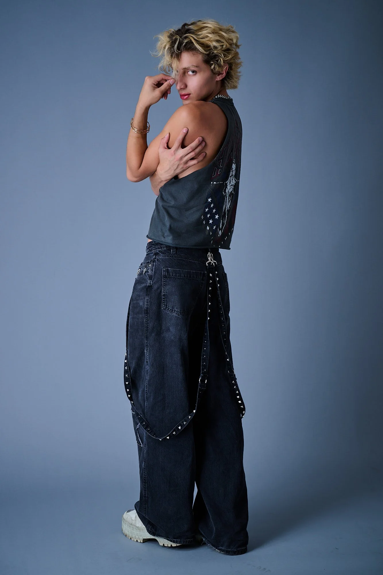 Person with curly blonde hair and fair skin standing against a gray background, wearing black baggy jeans, a sleeveless black graphic T-shirt, white sneakers, and gold bracelets, with one hand touching their face and the other hugging their shoulder.
