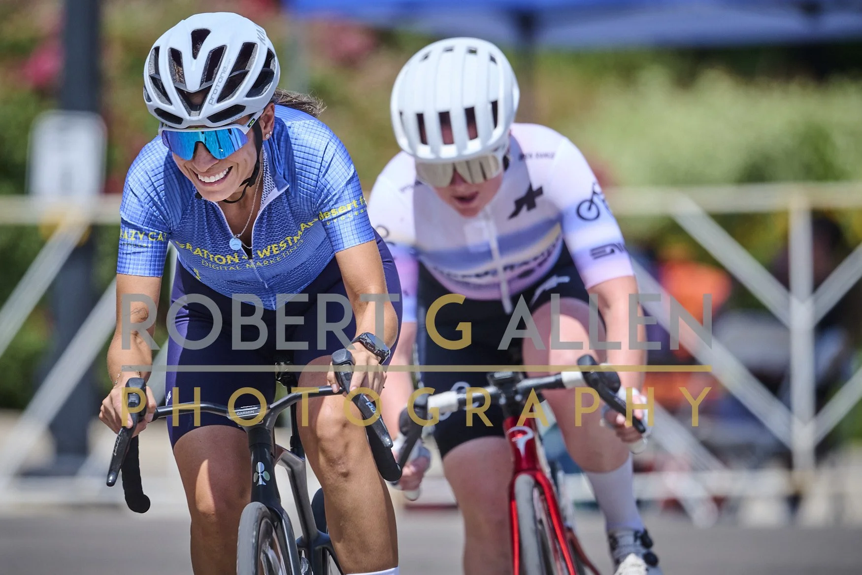 Two women wearing helmets and sunglasses riding bicycles side by side during a race or outdoor cycling event. They are smiling and appear focused. Background includes blurred trees and a metal fence.