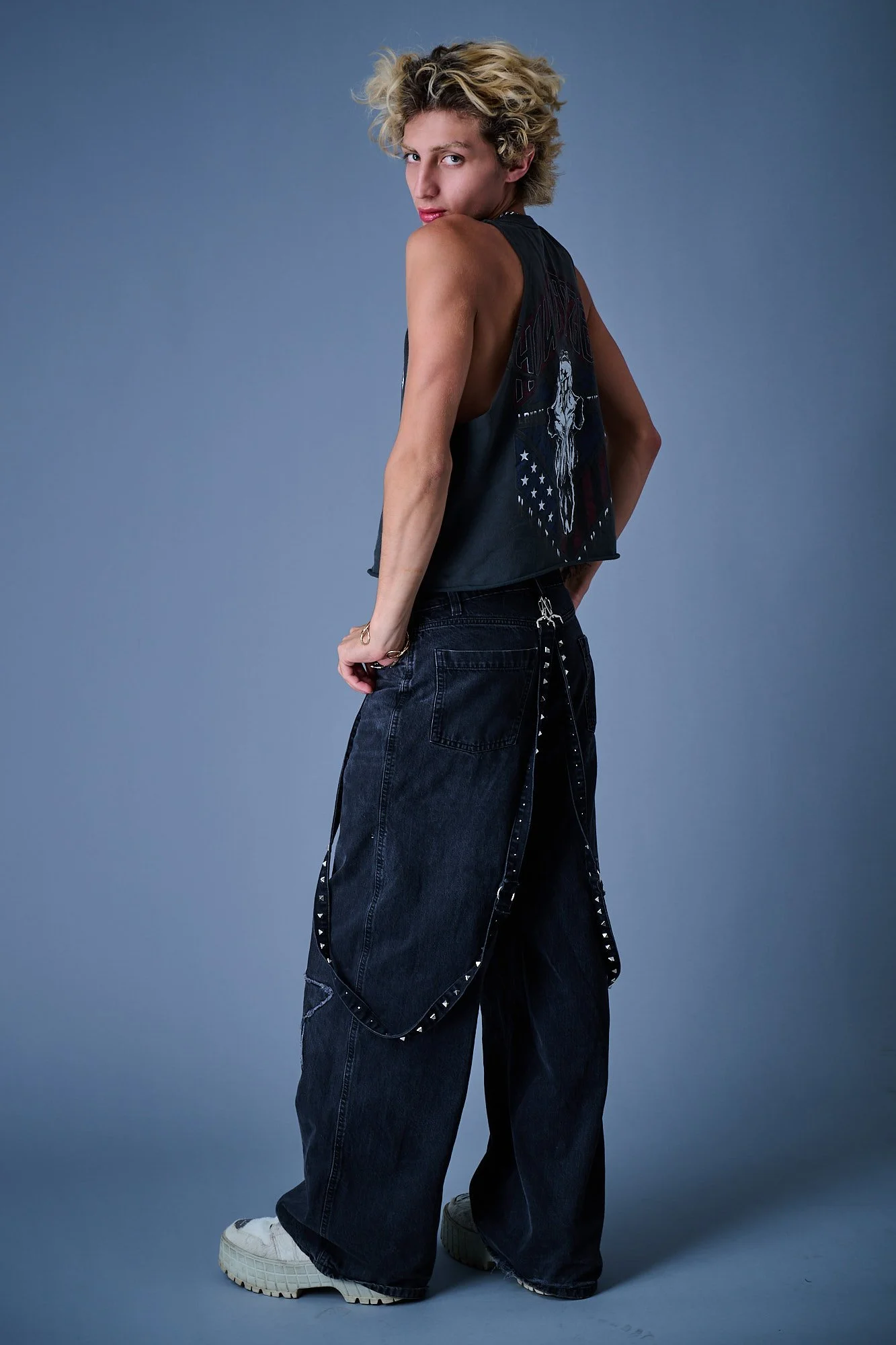 Person with curly blonde hair wearing a sleeveless graphic black top, baggy dark jeans with studded straps, and white sneakers, standing against a plain gray background.