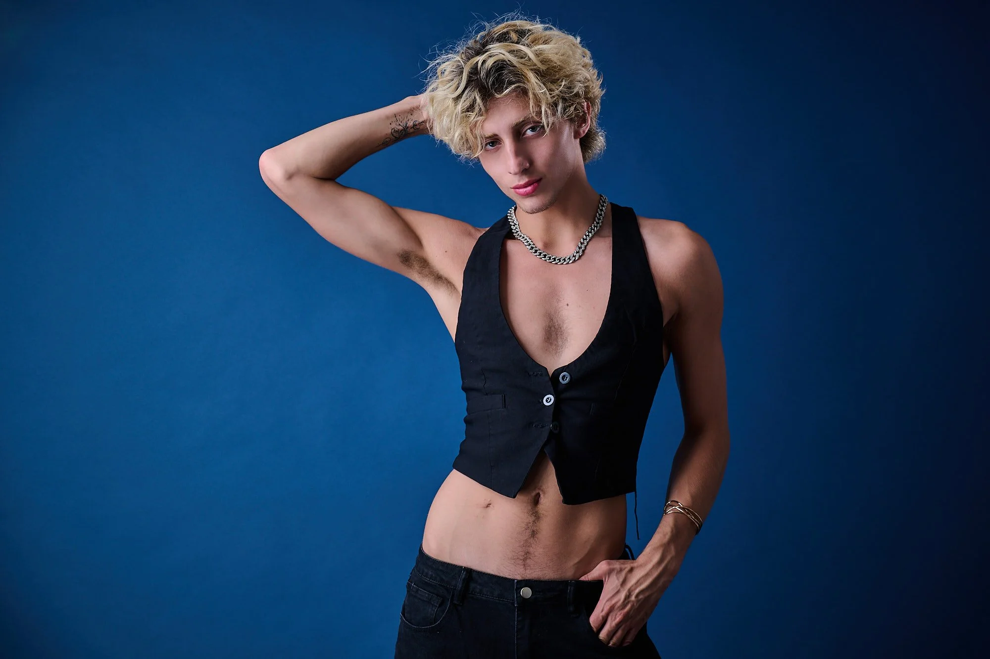 Young man with curly blonde hair, wearing a black sleeveless vest with buttons, a silver chain necklace, and dark jeans, standing against a blue background with one hand behind his head and the other on his hip.