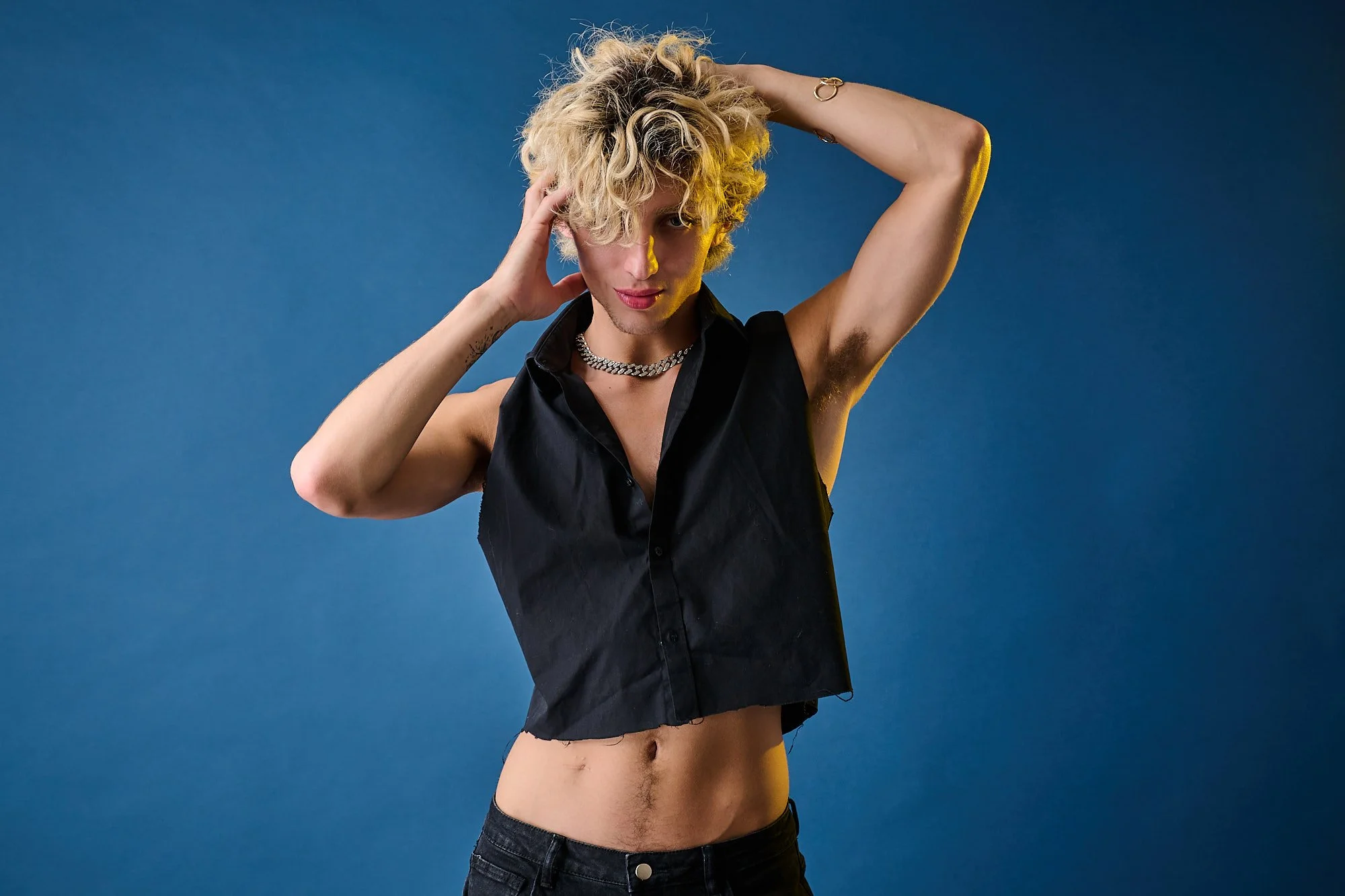 A person with curly blonde hair poses with one hand behind their head on a blue background, wearing a black sleeveless shirt, a silver chain necklace, and dark pants.