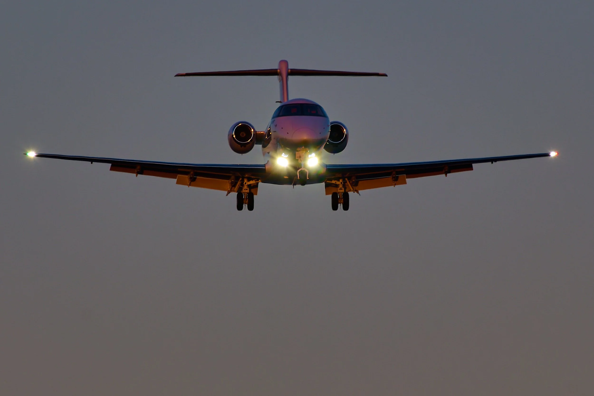 A commercial jet airplane flying in the sky at dusk with its headlights on and navigation lights on the wingtips visible.