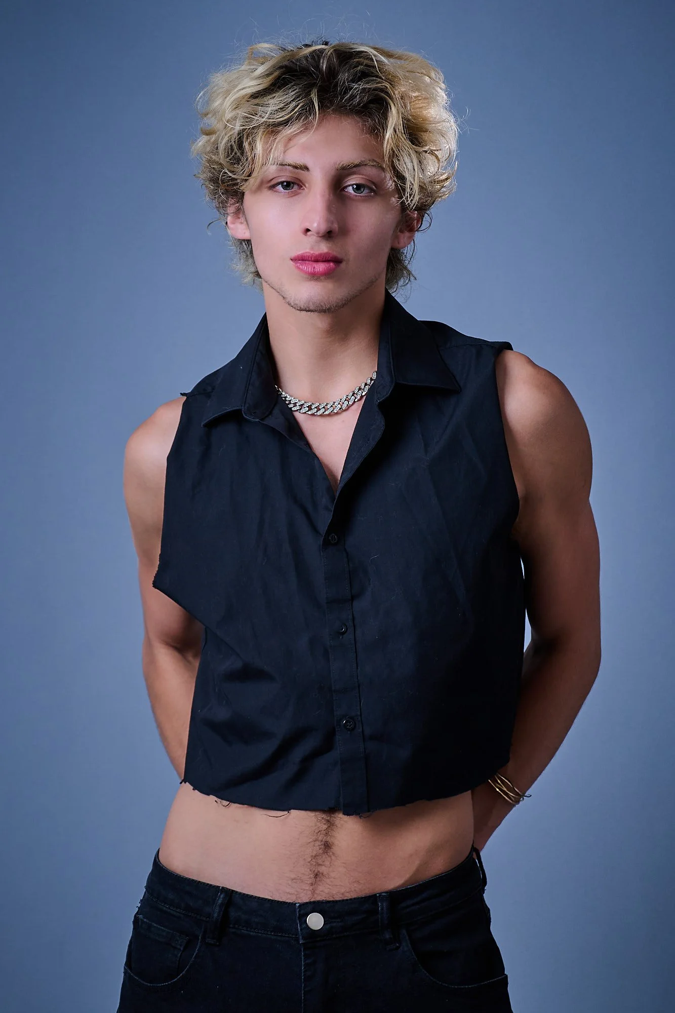A person with curly blond hair and choker necklace wearing a sleeveless black shirt with open bottom, standing against a plain blue background.