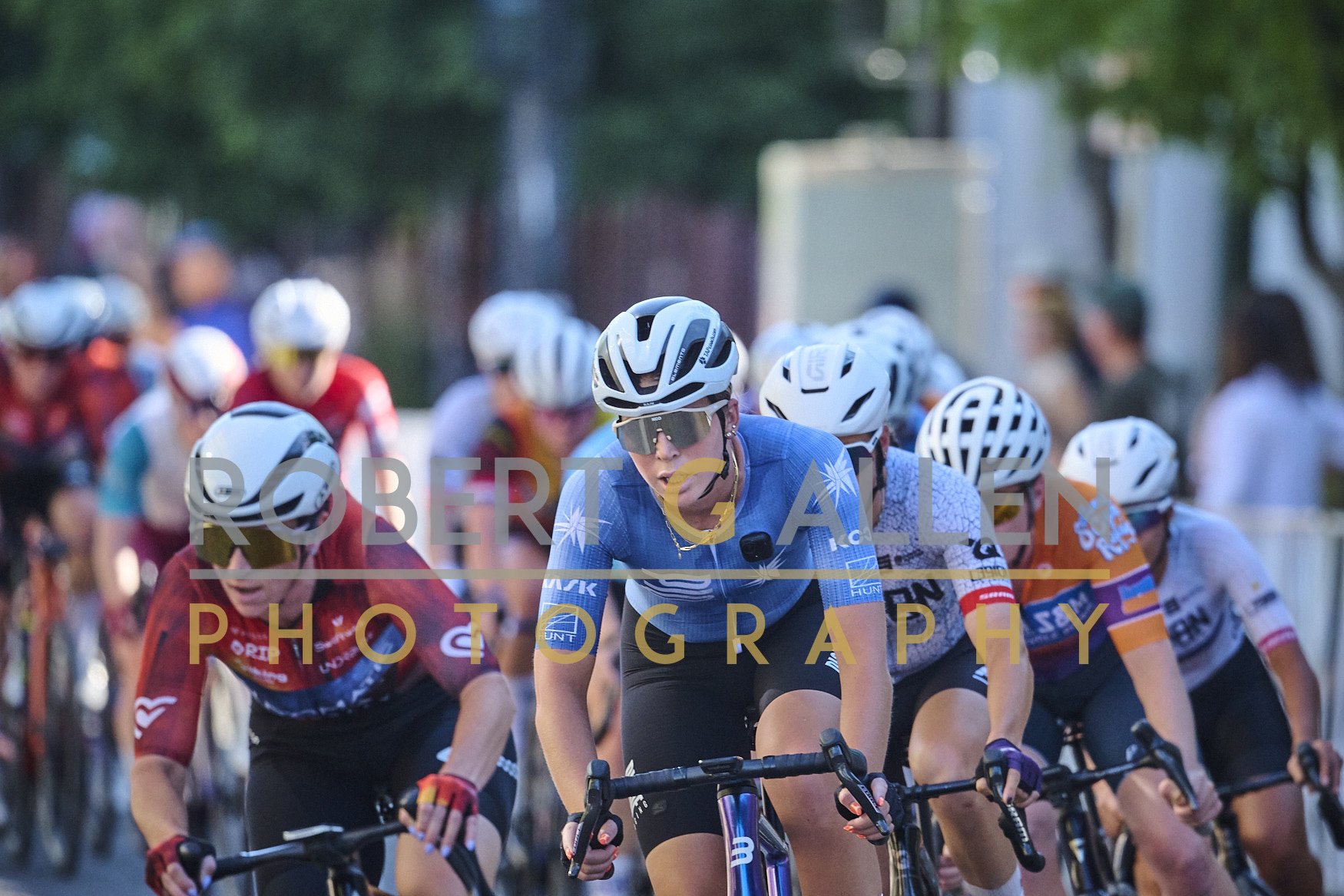 A group of cyclists riding in a race, with one cyclist in a blue jersey and sunglasses ahead of the others, all wearing helmets, in good outdoor lighting near trees and a fence.