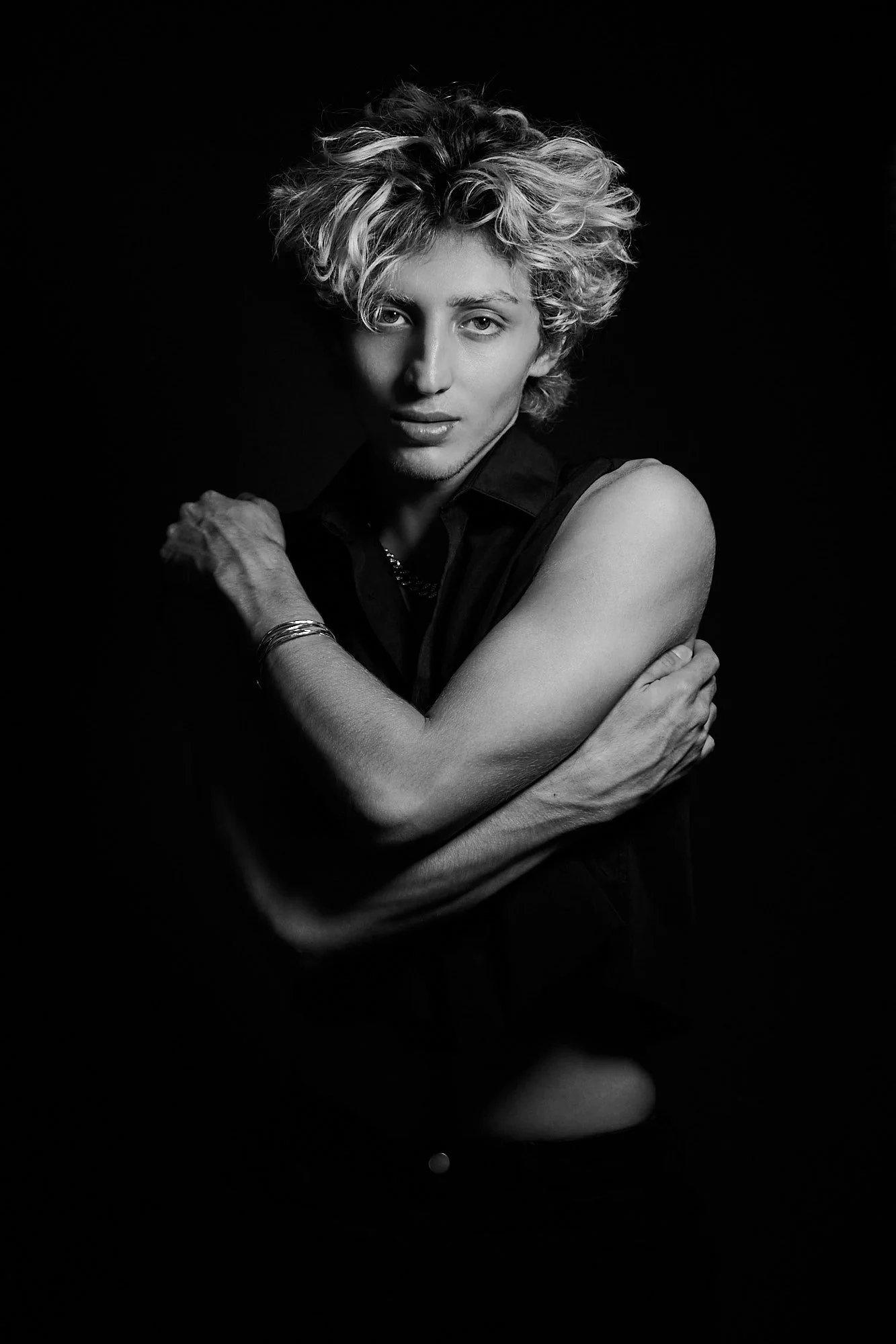 Black and white portrait of a person with curly hair, gazing at the camera, with their arms crossed, against a dark background.