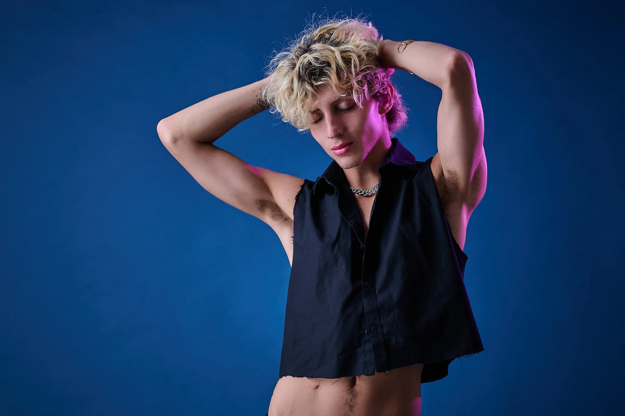 Young person with curly blond hair posing with hands behind head, wearing a black sleeveless shirt and silver chain necklace, against a solid blue background.