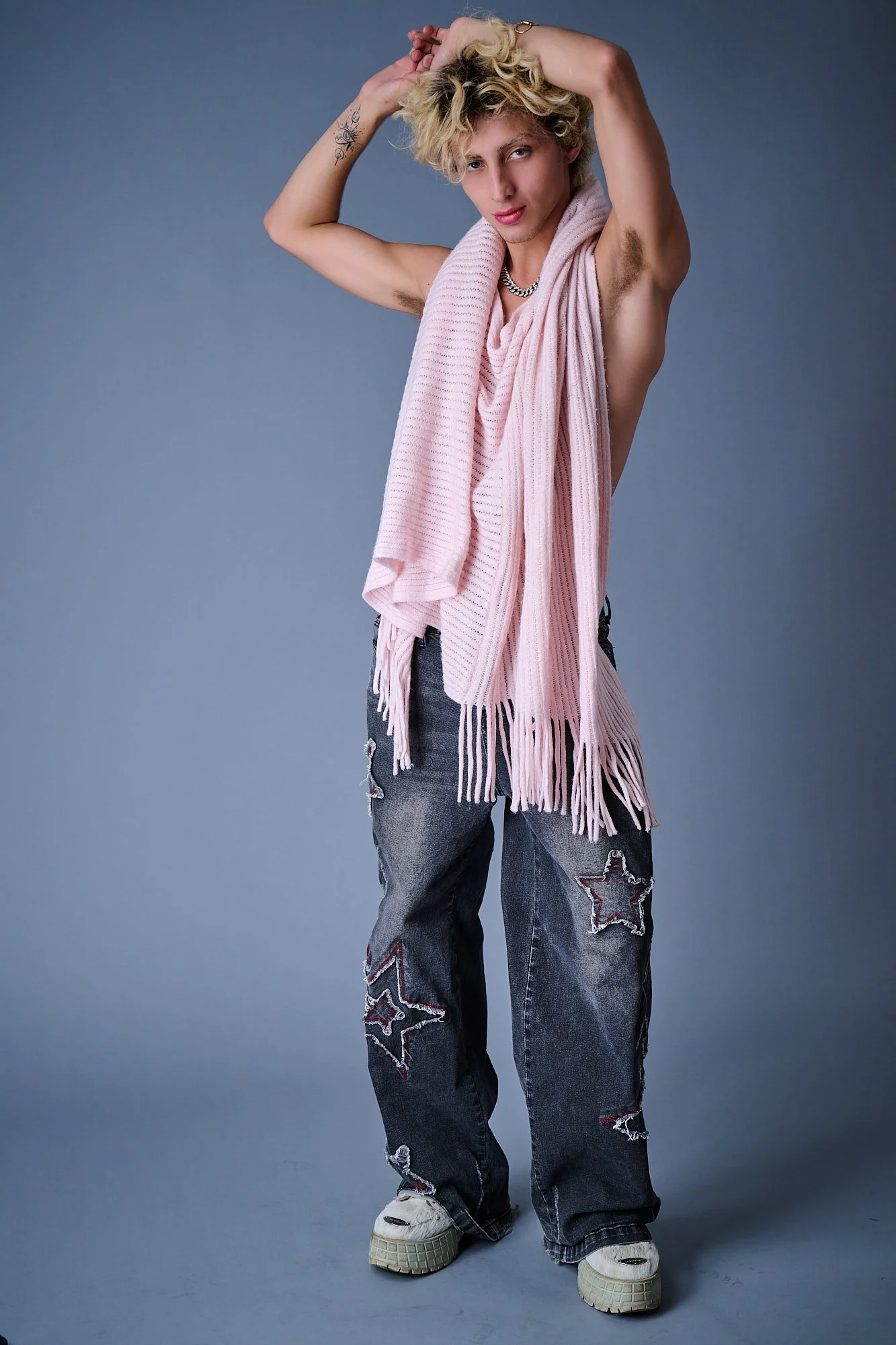 Young man with curly blond hair posing against a gray background, wearing distressed jeans with star patches, white sneakers, a chunky chain necklace, and a pink scarf draped over his shoulders, with one arm raised and his hand in his hair.