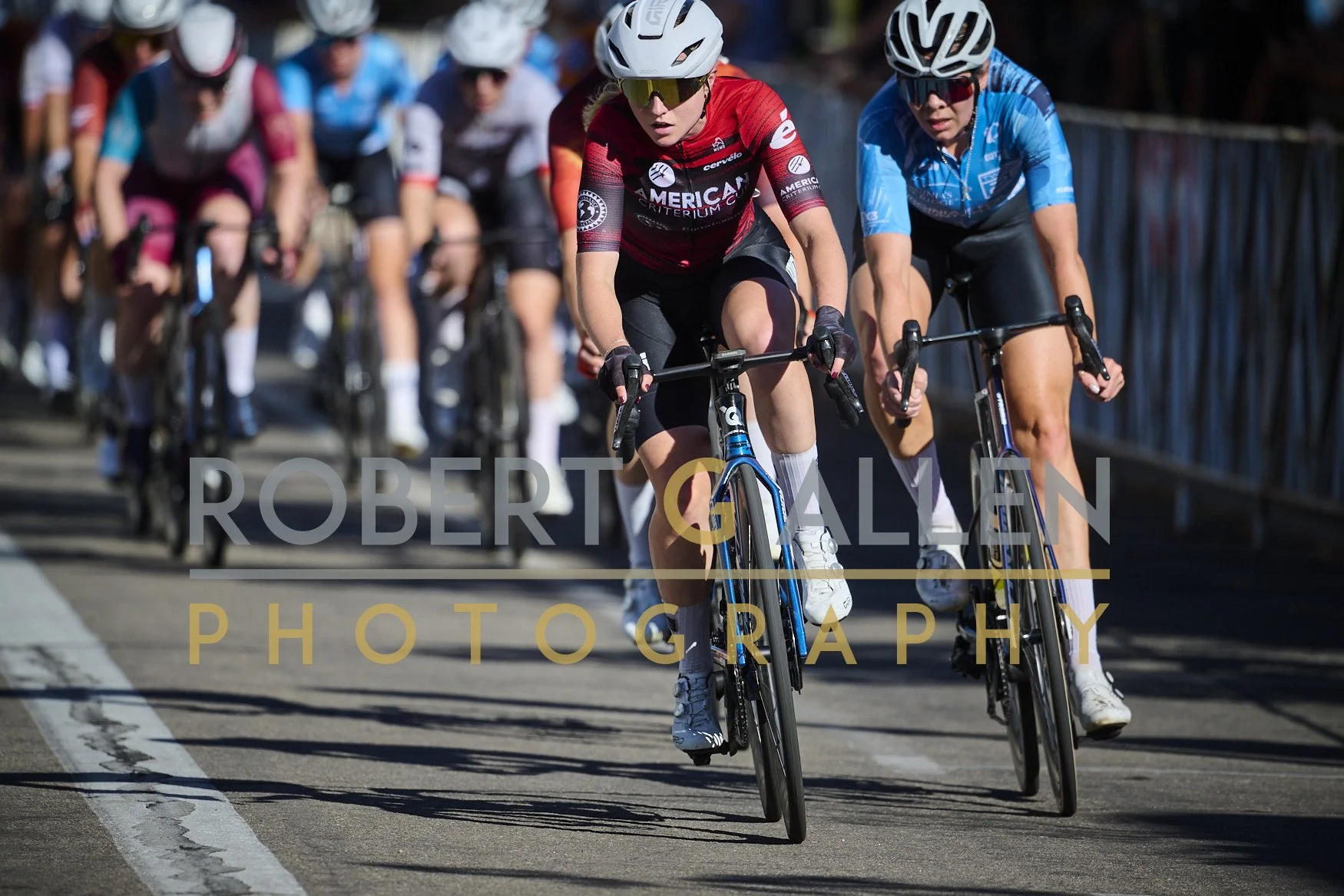 Cyclists participating in a race, with two women prominently in front wearing helmets and sunglasses, riding on a paved road with a railing on the side.