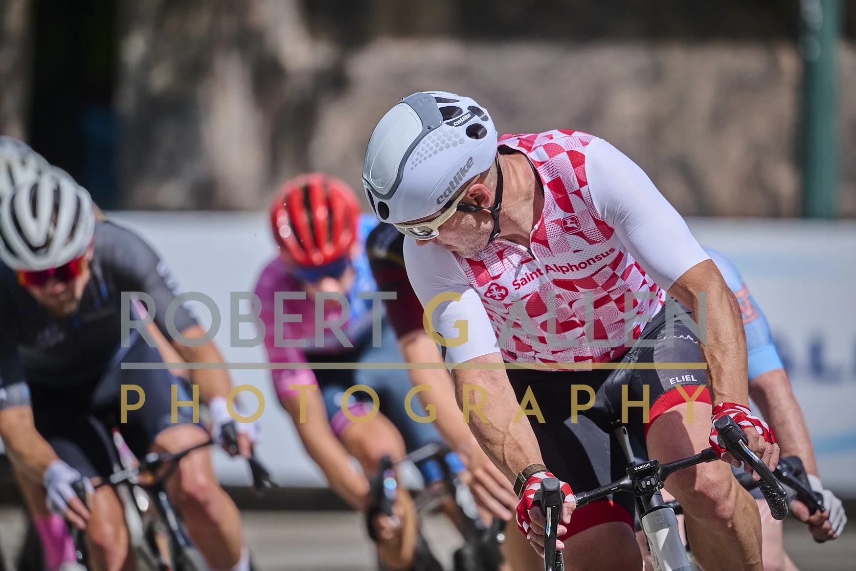 Cyclists participating in a race, with one cyclist wearing a white and red jersey, helmet, and sunglasses in the forefront, and others in colorful jerseys behind him.