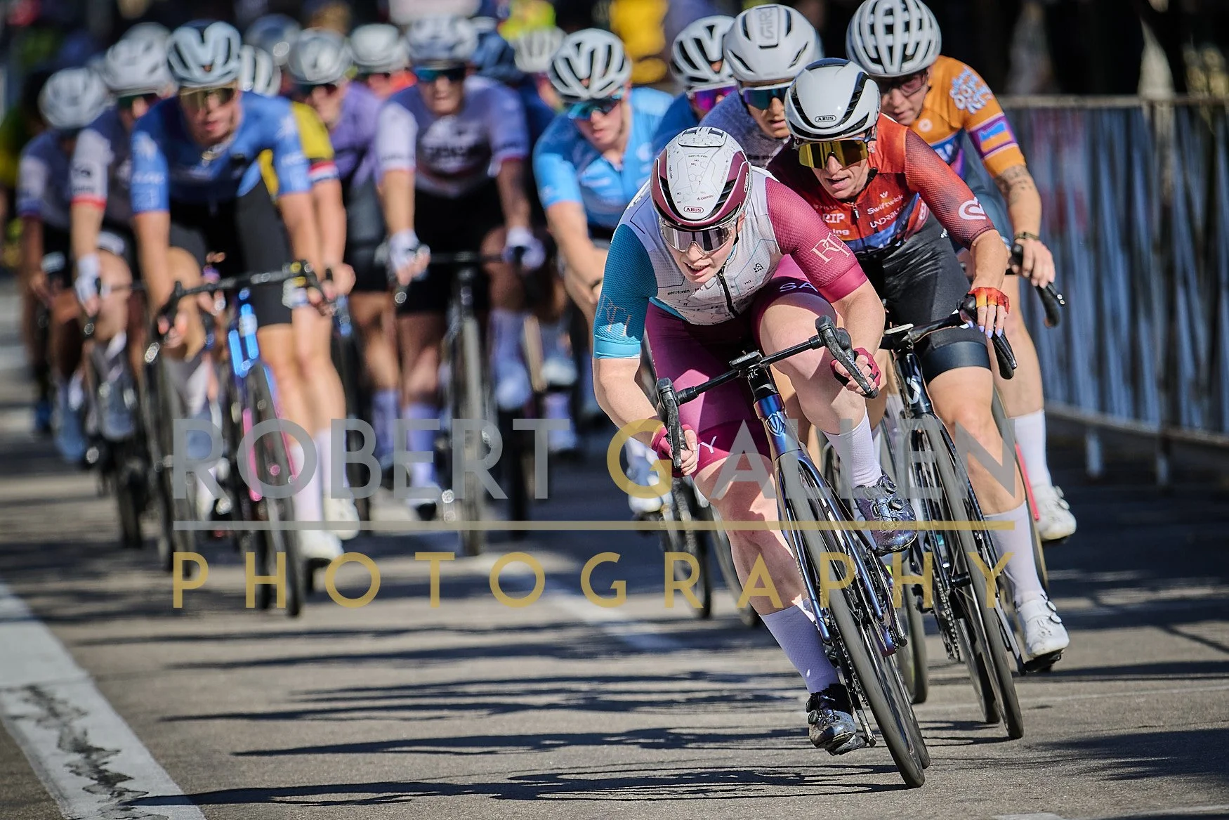 Group of women cyclists racing on a road, wearing helmets and colorful cycling outfits, with the lead cyclist in pink and white