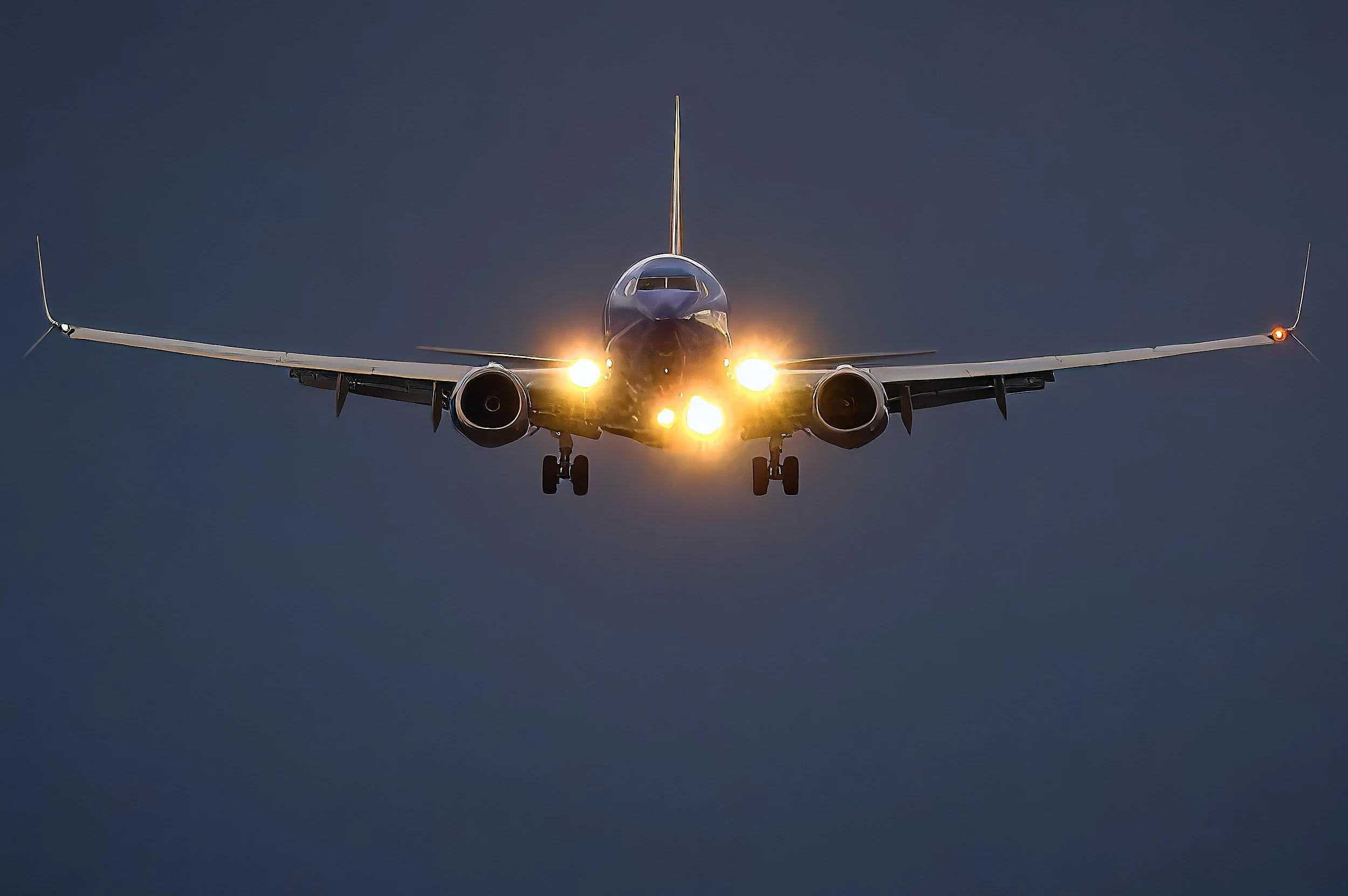 Passenger airplane in flight with its landing gear extended, flying against a cloudy evening sky with bright lights on the nose and engines.