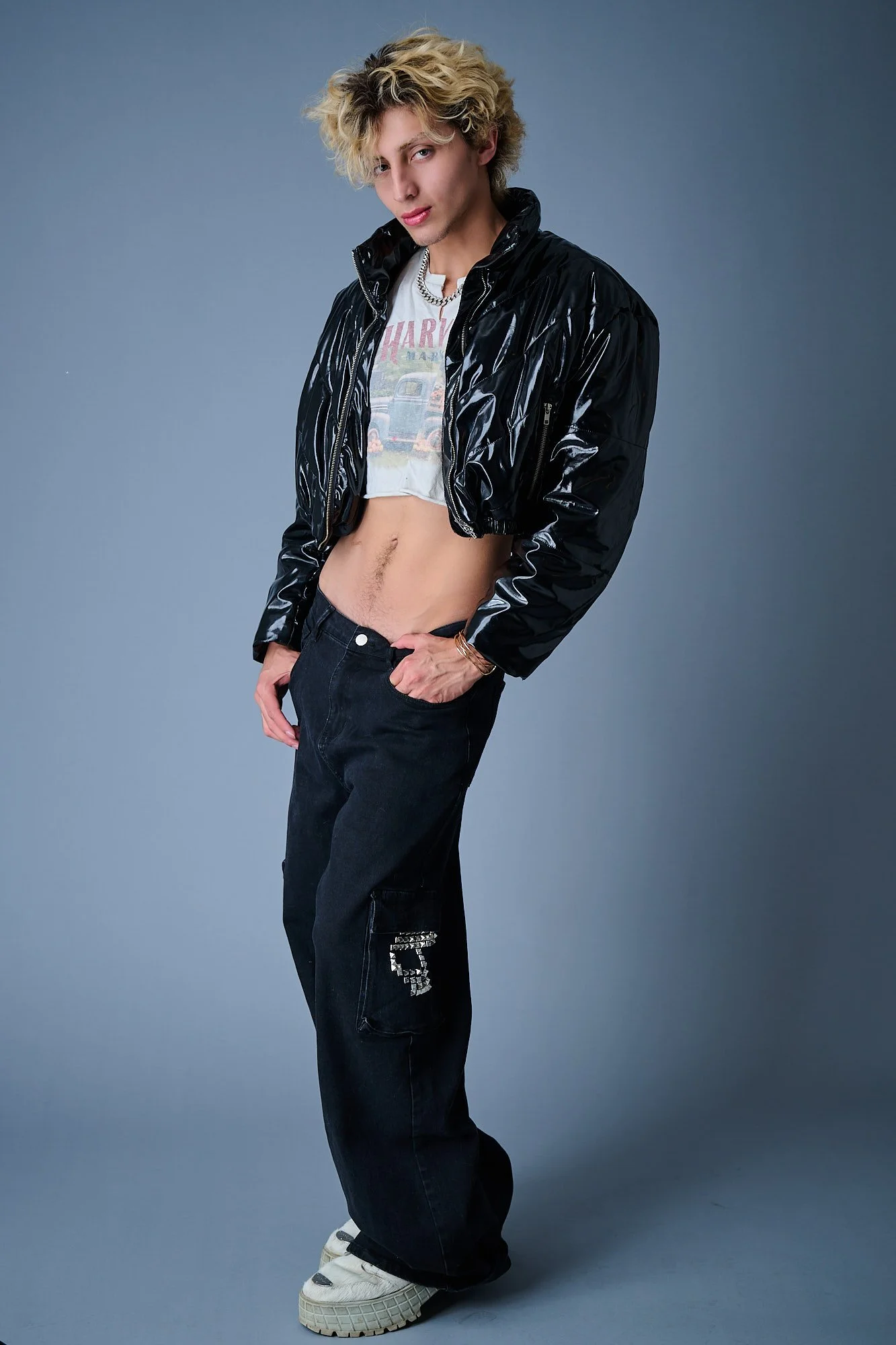 A young person with curly blond hair wearing a shiny black jacket, a graphic T-shirt, baggy dark jeans with a patch on the leg, and white sneakers, posing with one hand in their pocket against a plain gray background.