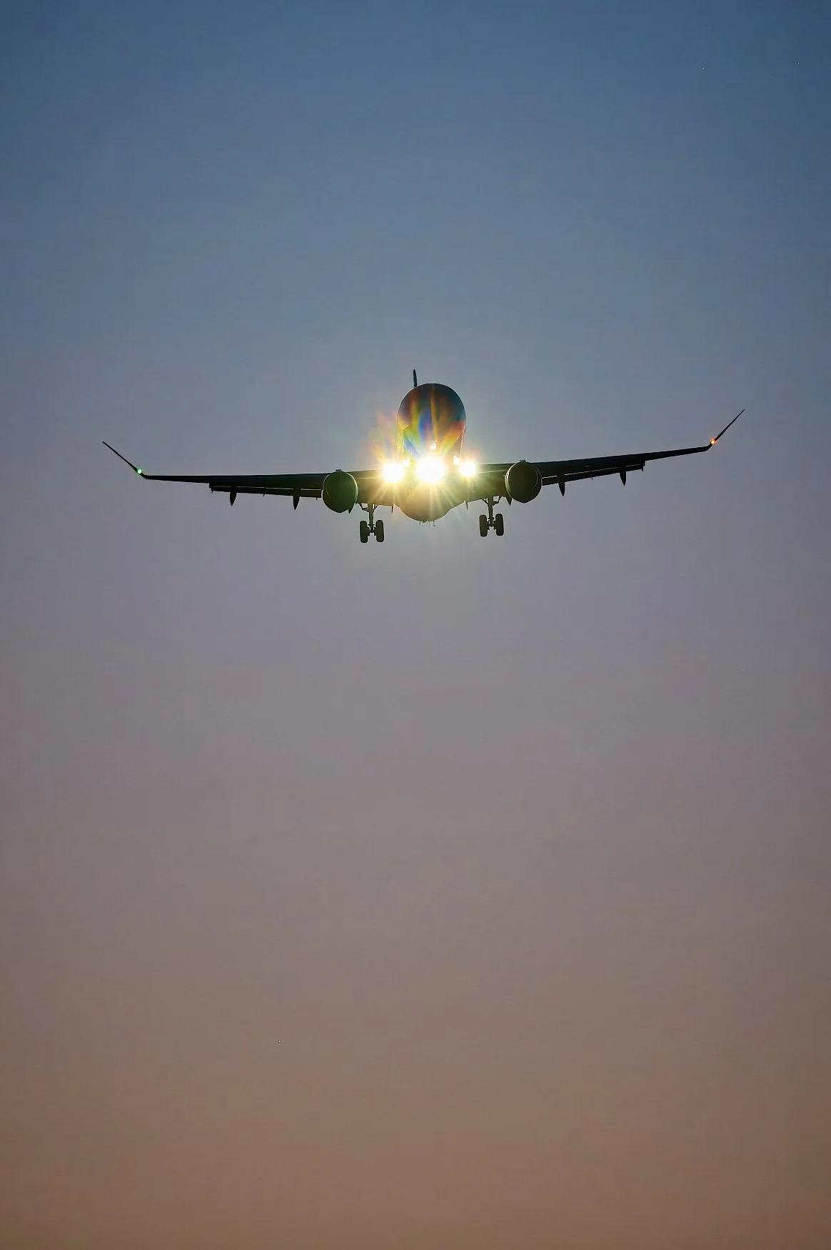An airplane flying with its lights on against a dusky sky.