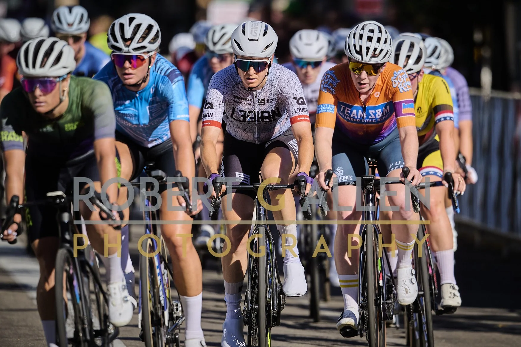 A group of cyclists riding in a race or event, wearing colorful jerseys and helmets, with some using sunglasses, on a sunny day.