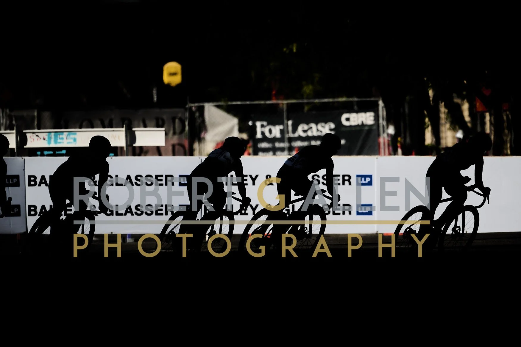 Silhouettes of four cyclists riding their bikes at night during a cycling event, with banners and signs in the background.