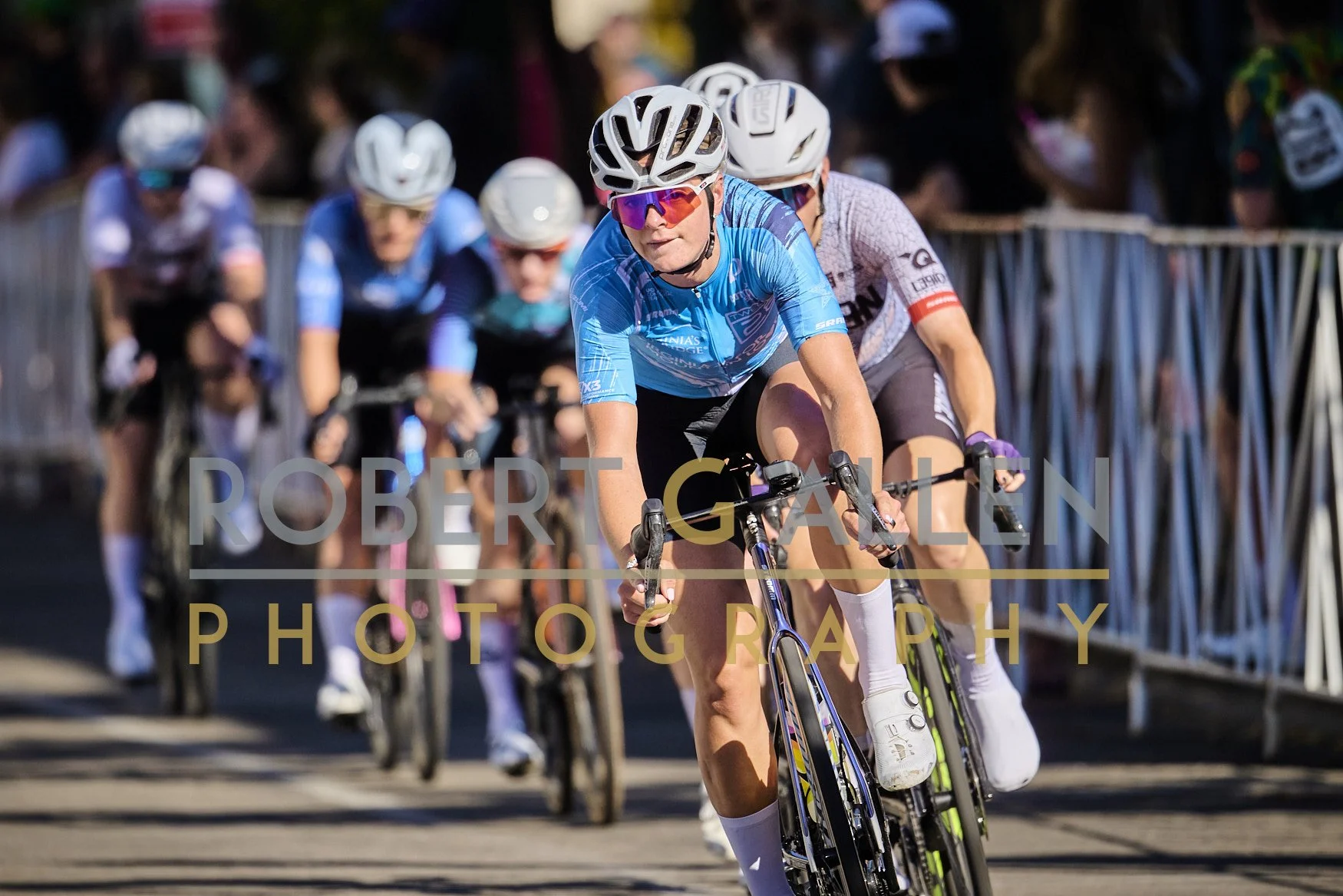Cyclists in a race with a crowd watching behind metal barriers, focusing on a woman in blue jersey and black shorts wearing a white helmet and sunglasses, riding a racing bike.
