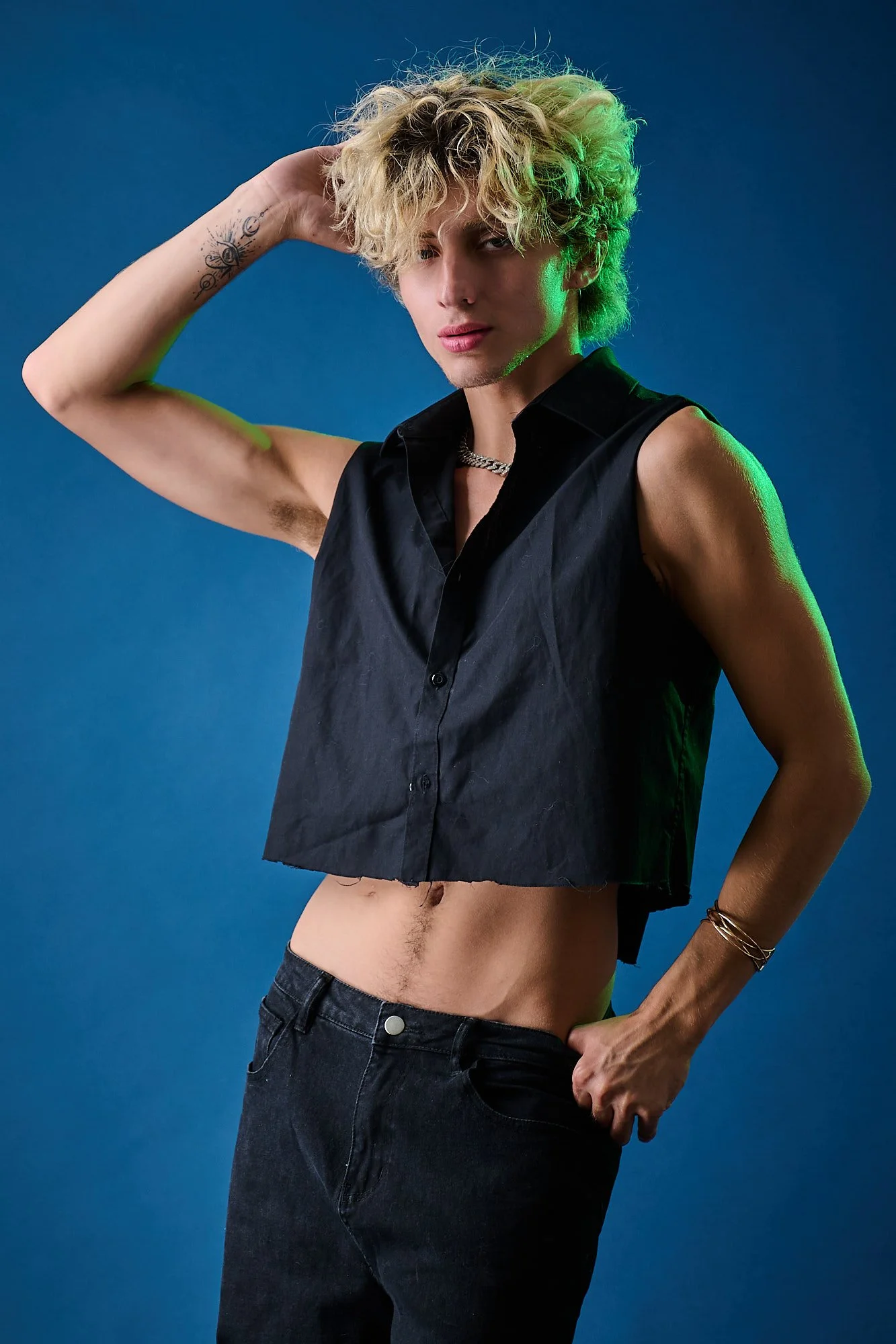A young man with blonde, curly hair pose shirtless with a black sleeveless shirt, dark denim jeans, against a blue background. He has tattoos on his left forearm and is wearing jewelry, including a chain necklace and bracelets.