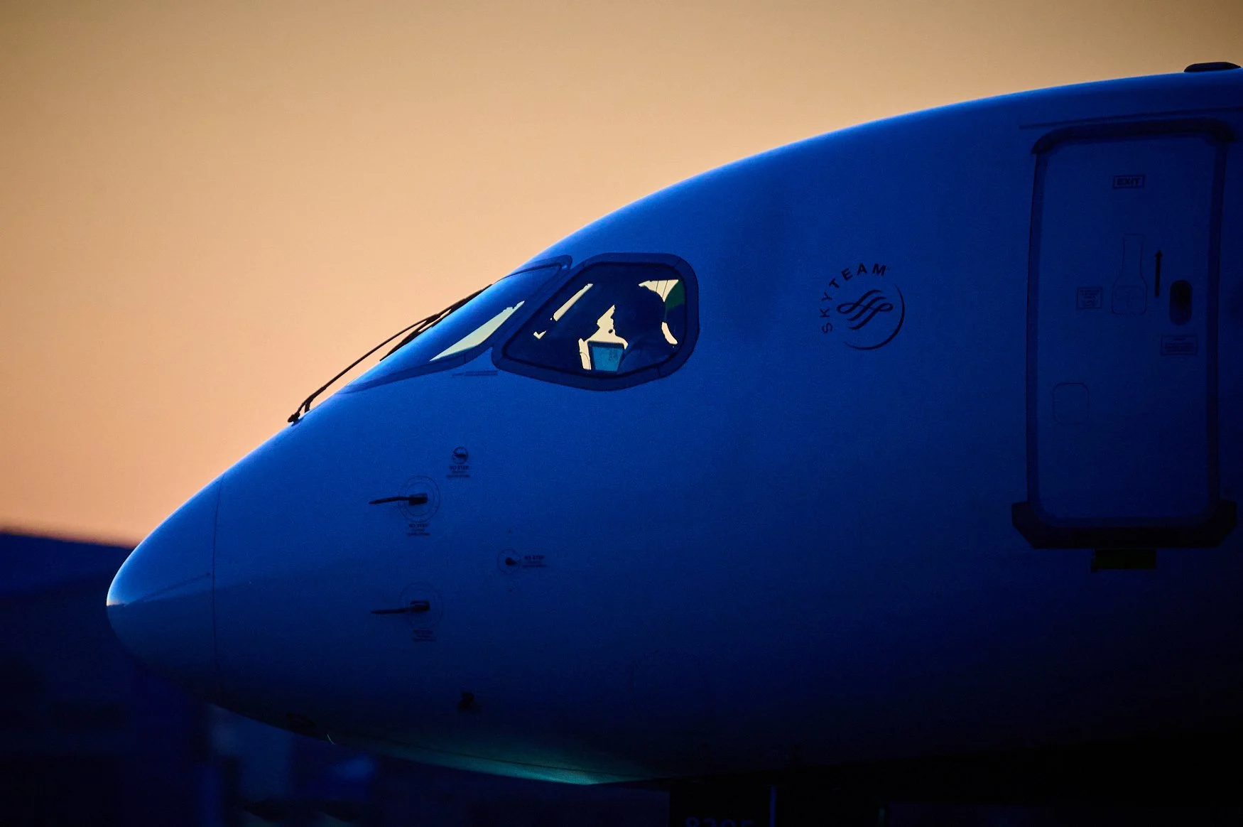 Close-up of a commercial airplane's cockpit with a silhouette of a person inside, set against a soft gradient sky at dusk.