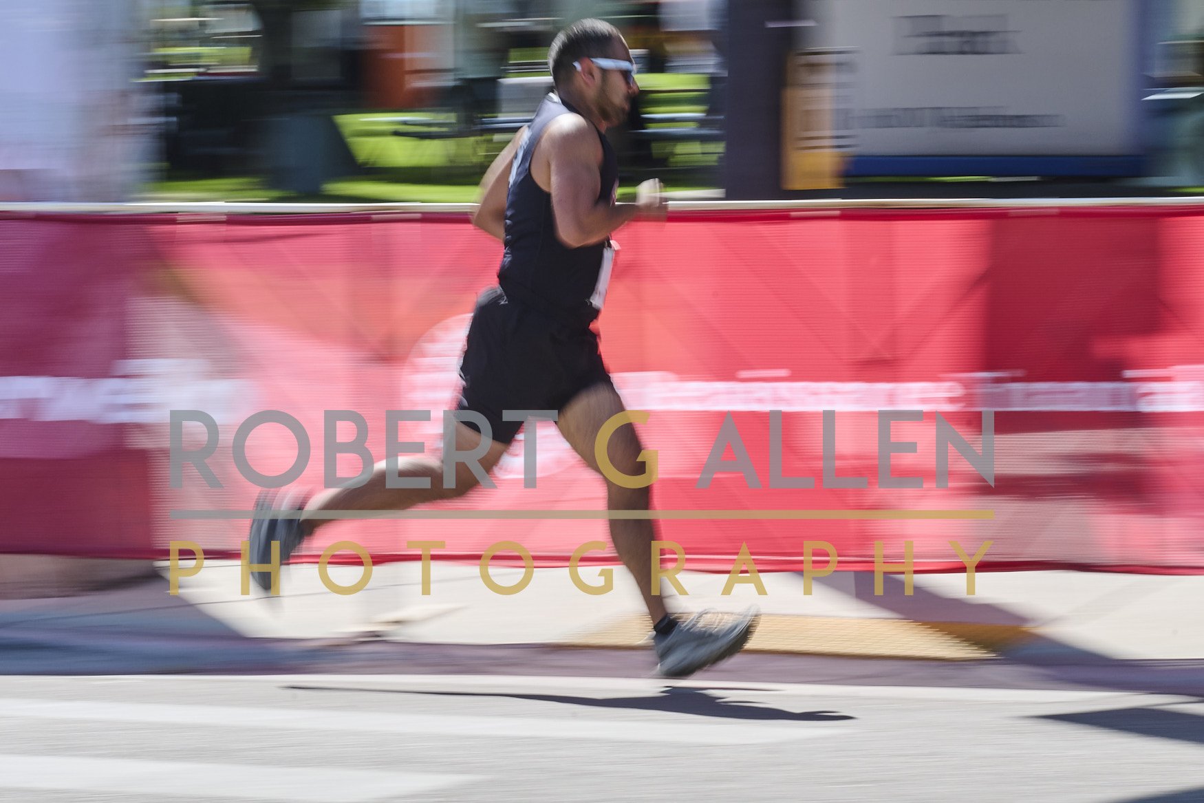 A male runner in athletic gear and sunglasses running on a city street during a race, with motion blur background and a red barrier with advertising, and the text 'ROBERT G ALLEN PHOTOGRAPHY' overlaid.