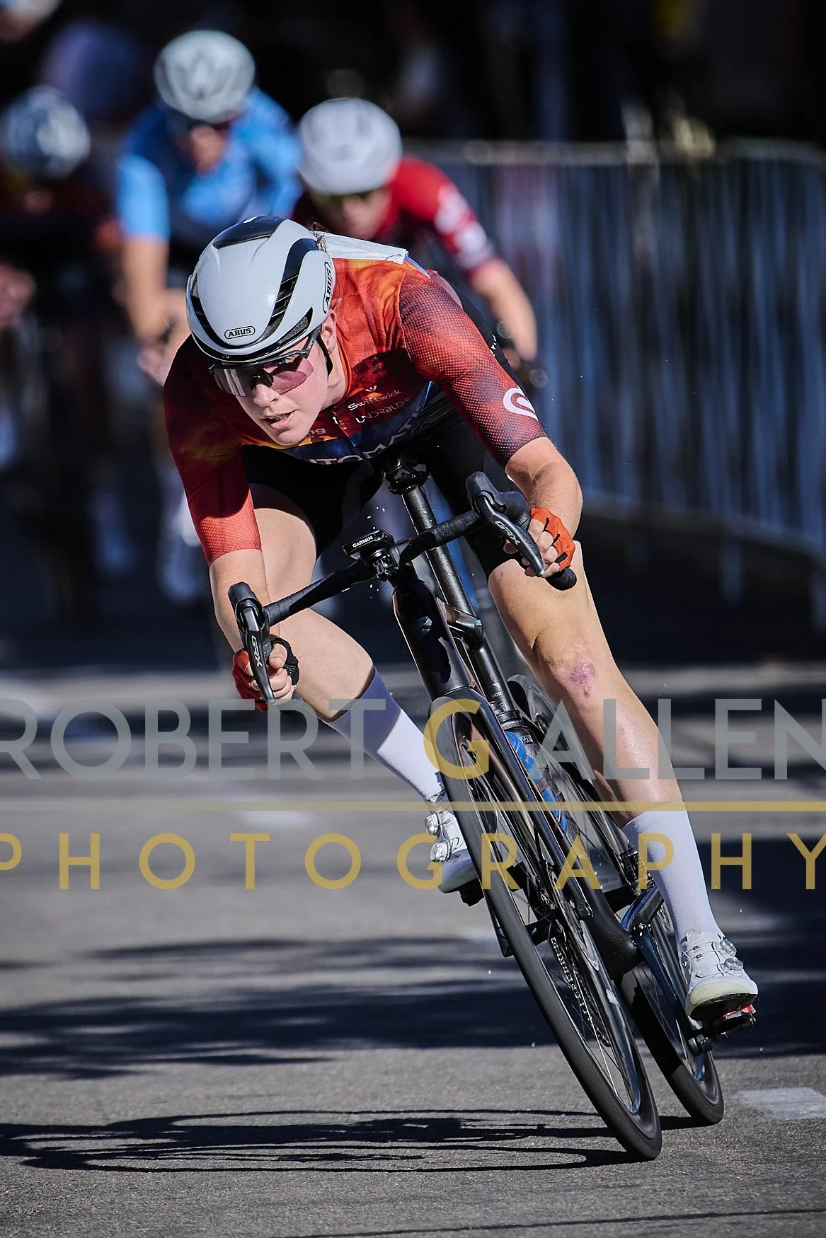 Cyclist in a red and black jersey leaning into a turn on a road race bike, with a bruise or injury on his leg, wearing a white helmet and sunglasses, with background spectators.