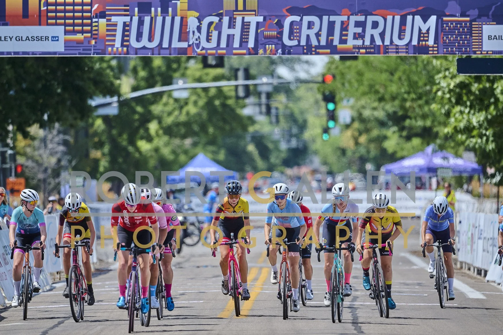 A group of cyclists participating in a race, wearing colorful jerseys and helmets, riding on a city street marked with yellow lines, with trees and event banners in the background.