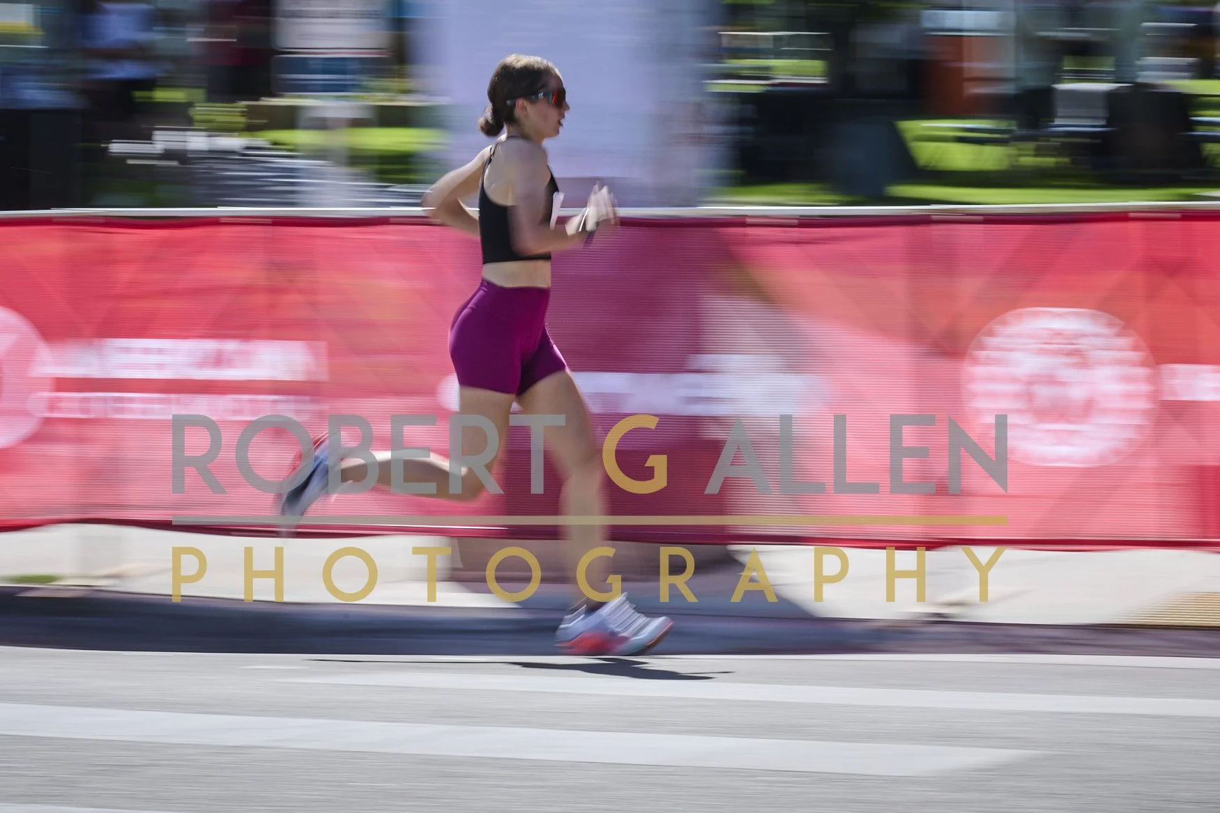 A female runner dressed in black and purple running gear, wearing sunglasses, running along a city street with a blurred background and a red barrier with the text 'ROBERT GALLEN PHOTOGRAPHY' overlaid.