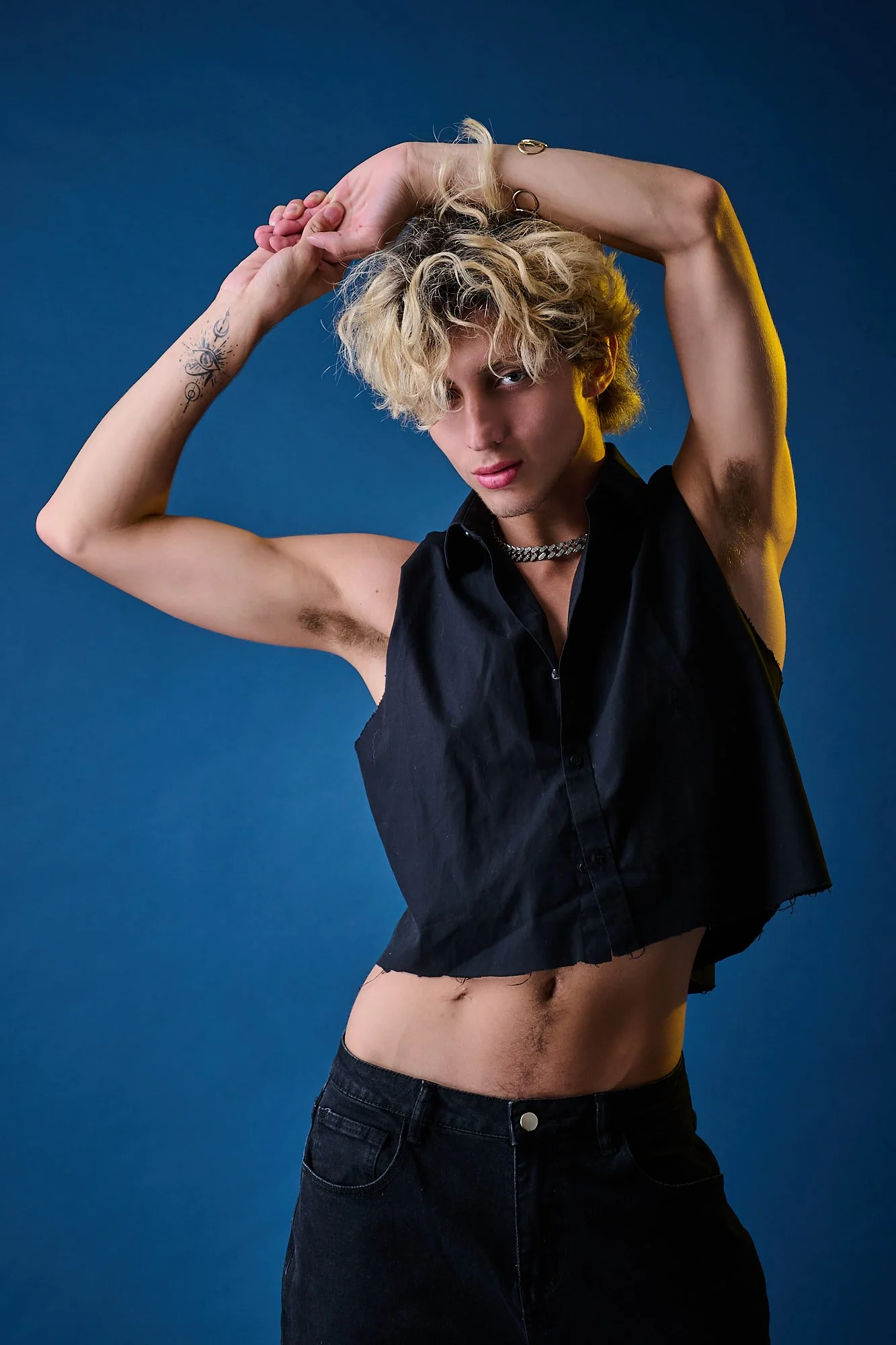 A young person with blonde curly hair posing against a blue background. They are wearing a sleeveless black shirt, black jeans, a silver chain necklace, and have tattoos on their arms. Their left arm is raised, resting on their head, and they are loo