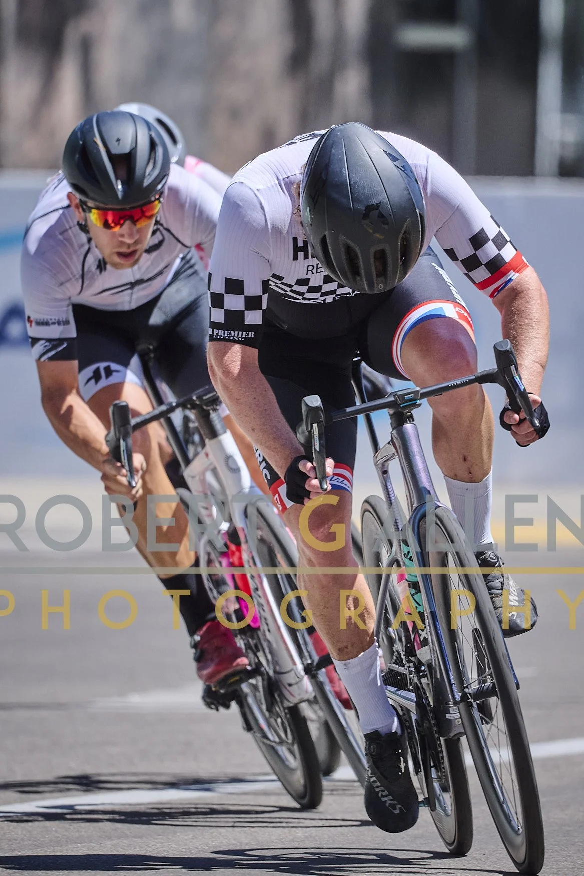 Two cyclists in a race leaning into a turn on a road, wearing helmets and sports gear.