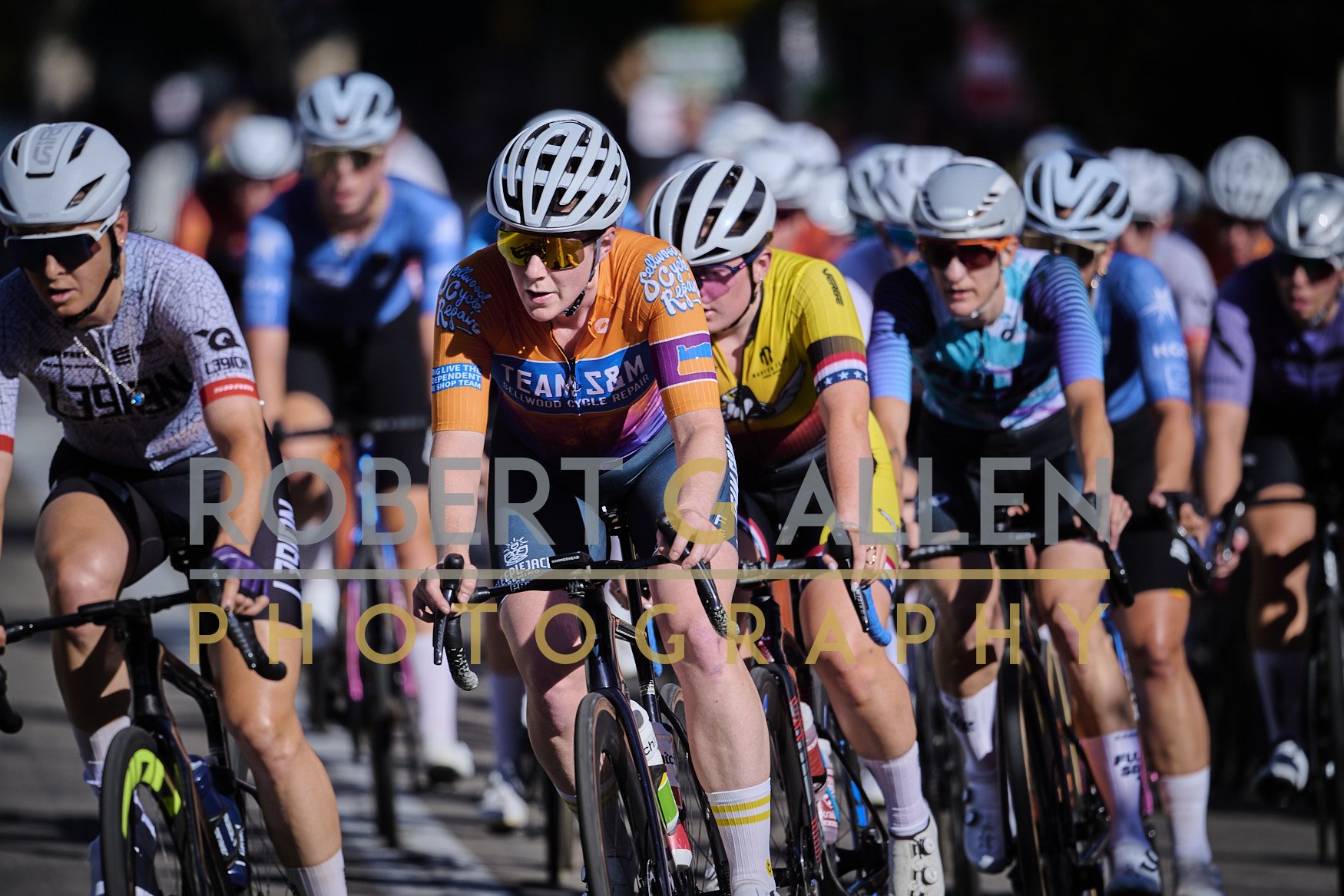Cyclists wearing helmets and colorful jerseys participate in a race, riding closely together on a paved road.