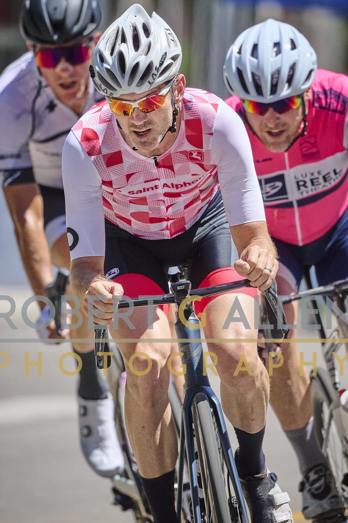 Three men wearing cycling helmets and sunglasses participating in a bike race, heavily focusing on the man in the middle in a red and white jersey. The background is outdoors with a fence.