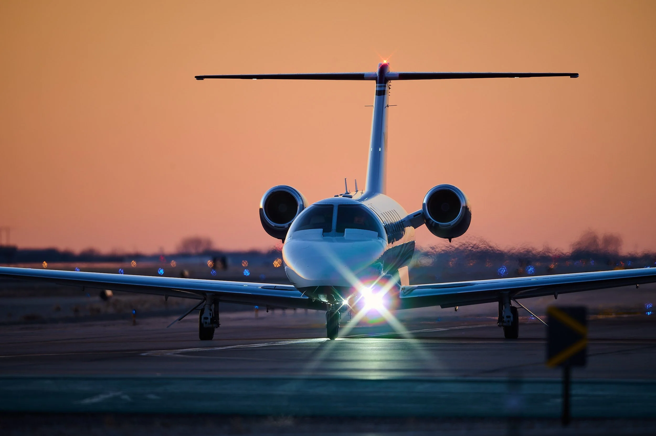 A private jet on the runway at sunset, viewed from the front with a clear sky and warm orange hues in the background.