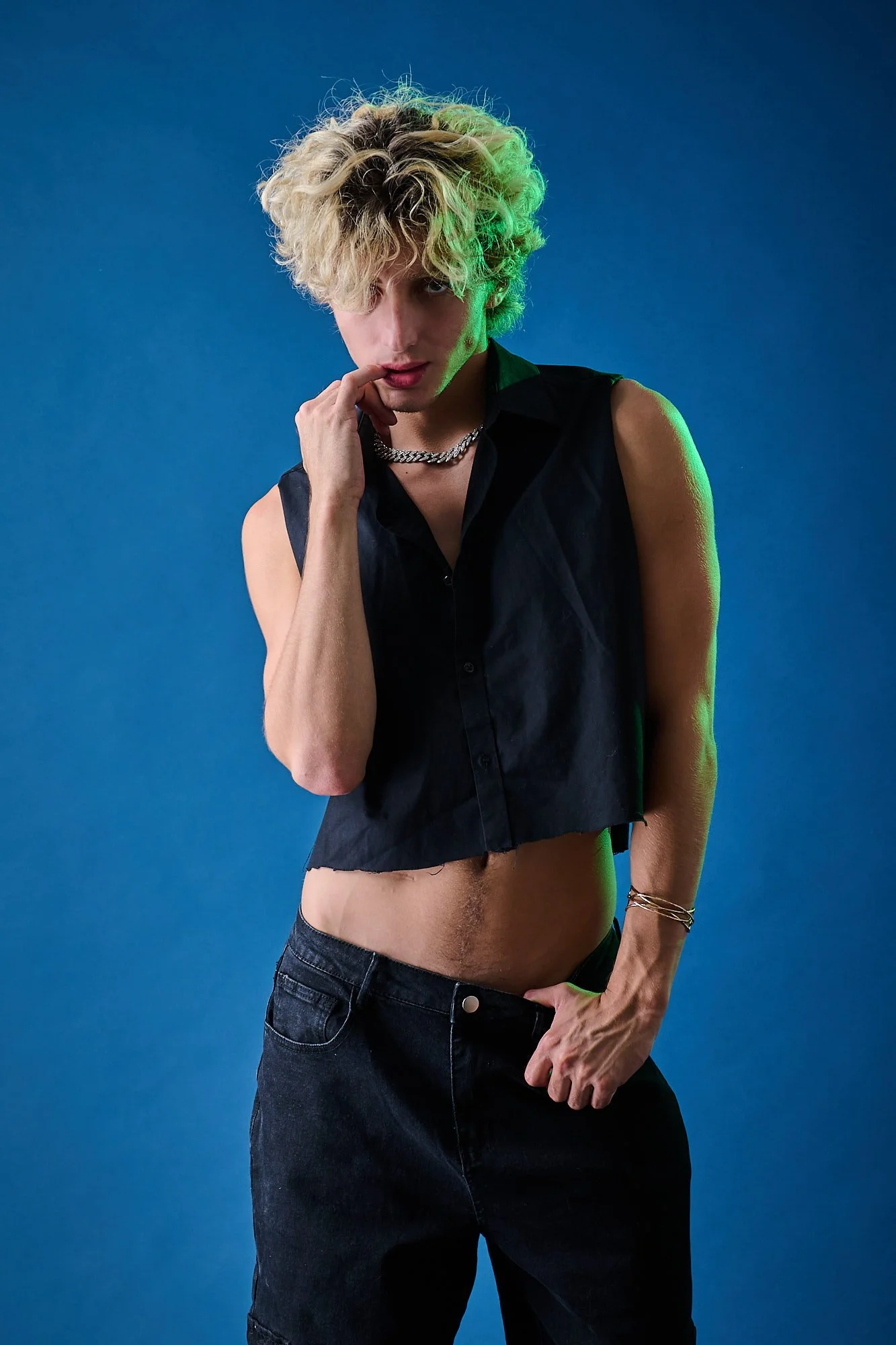 Person with curly blonde hair wearing a sleeveless black shirt, dark jeans, and jewelry against a blue background.