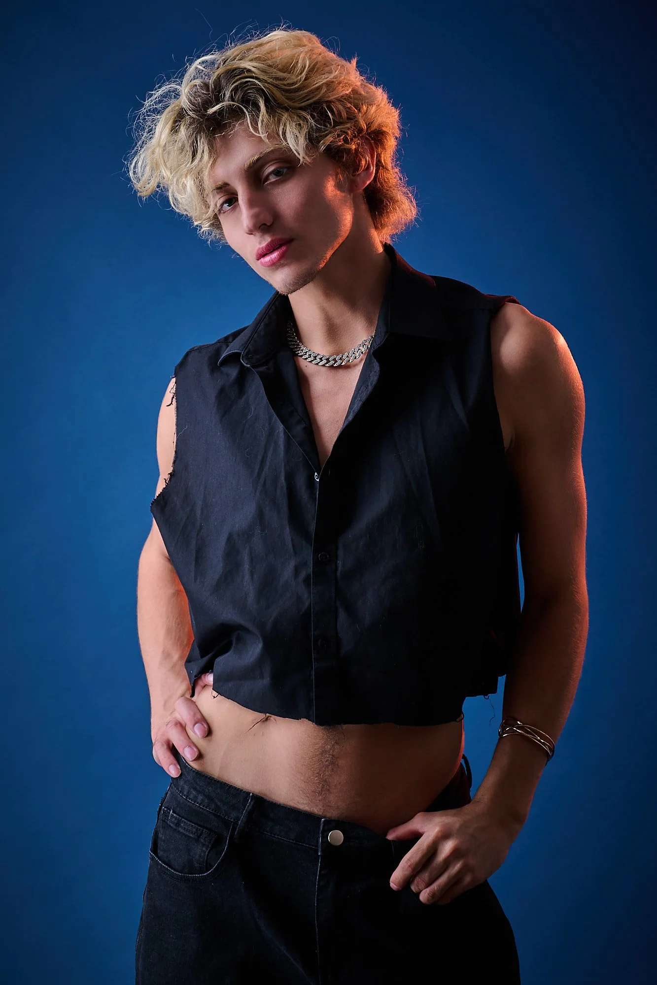 A person with light skin, curly blonde hair, and makeup posing against a blue background. They are wearing a sleeveless black shirt with some parts torn, a silver chain necklace, and black jeans. Their expression is confident with slightly parted lip