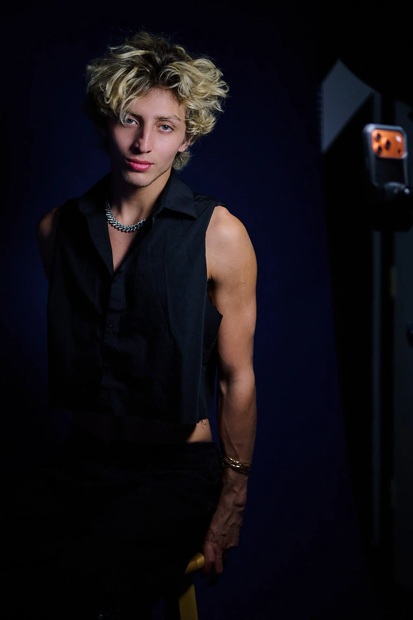 A person with short blond curly hair, wearing a black sleeveless shirt, a chain necklace, and bracelets, standing in front of a dark background with a red and black camera on a stand visible in the background.