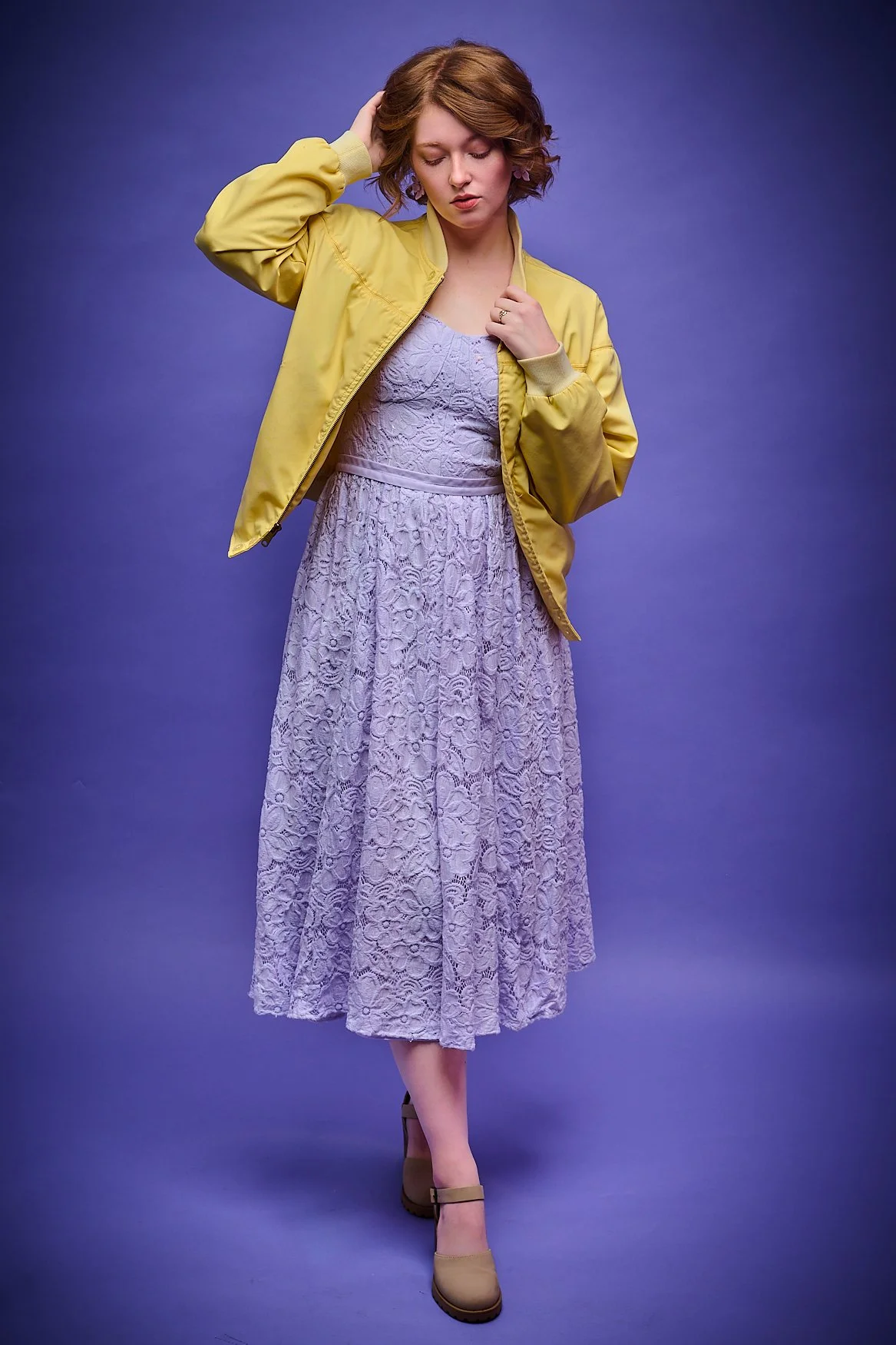 A young woman with short curly red hair wearing a lavender lace dress and a yellow jacket against a purple background.