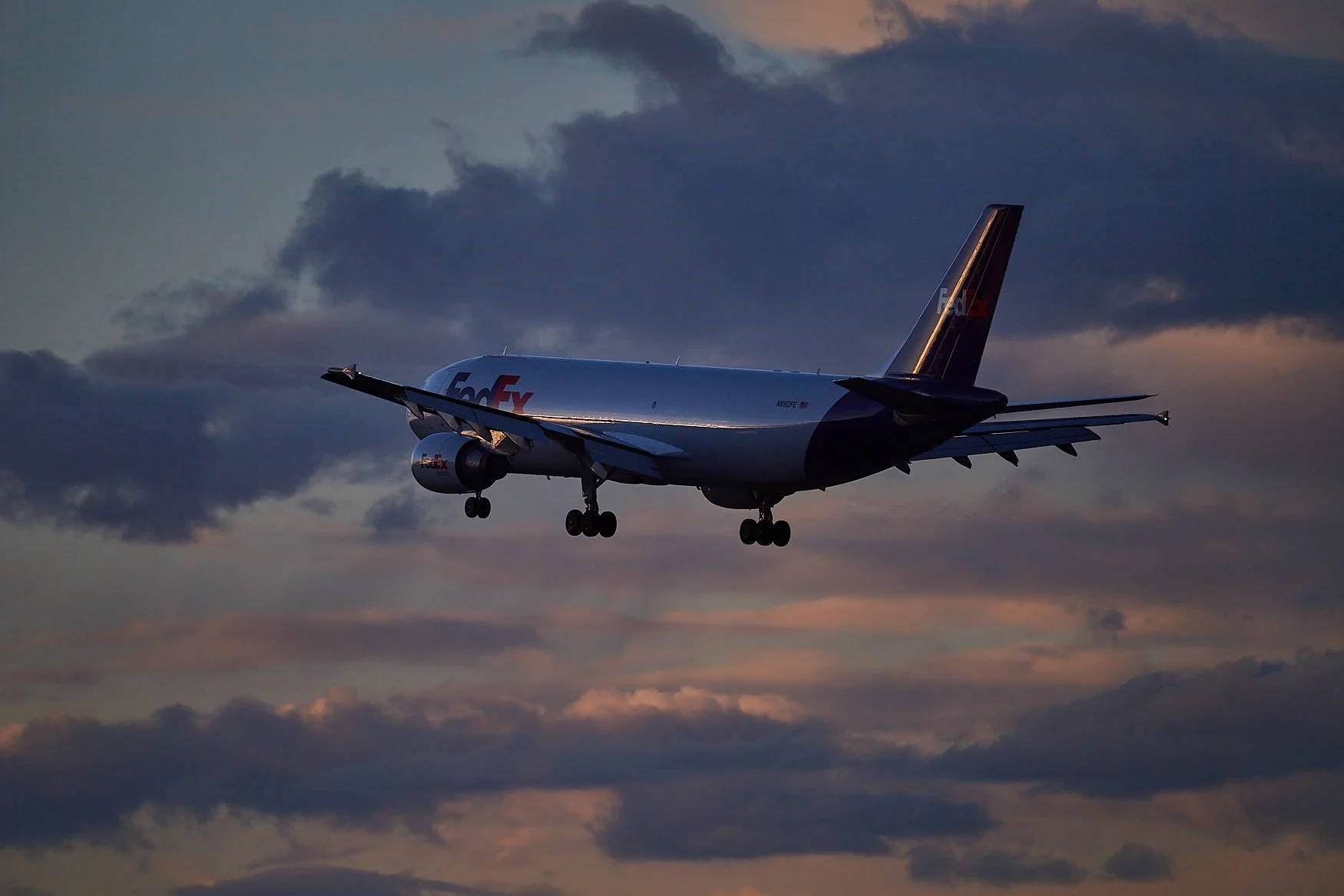 A FedEx cargo airplane flying in the sky during sunset, with dark clouds and a colorful sky in the background.