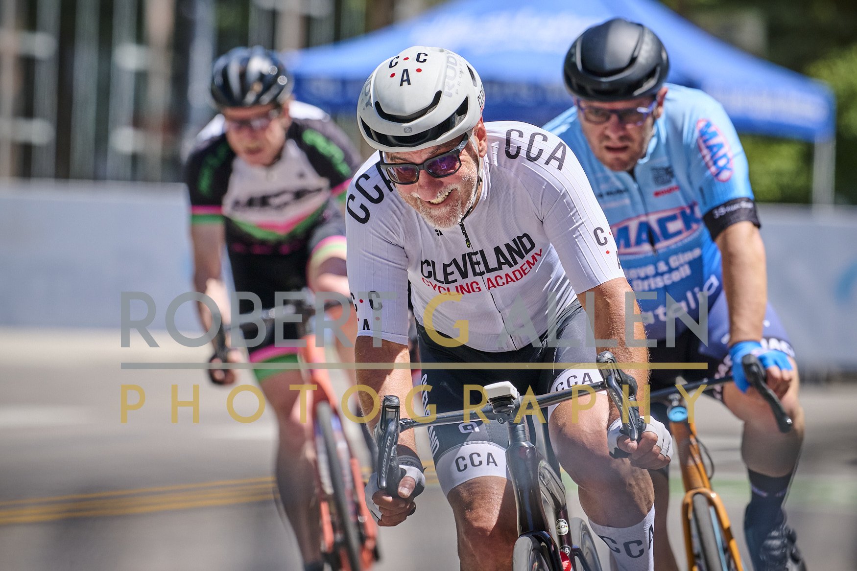 Group of senior cyclists riding uphill during a race or event, wearing branded cycling jerseys and helmets, with the central focus on a smiling man in a white Cleveland Cycling Academy jersey and helmet.