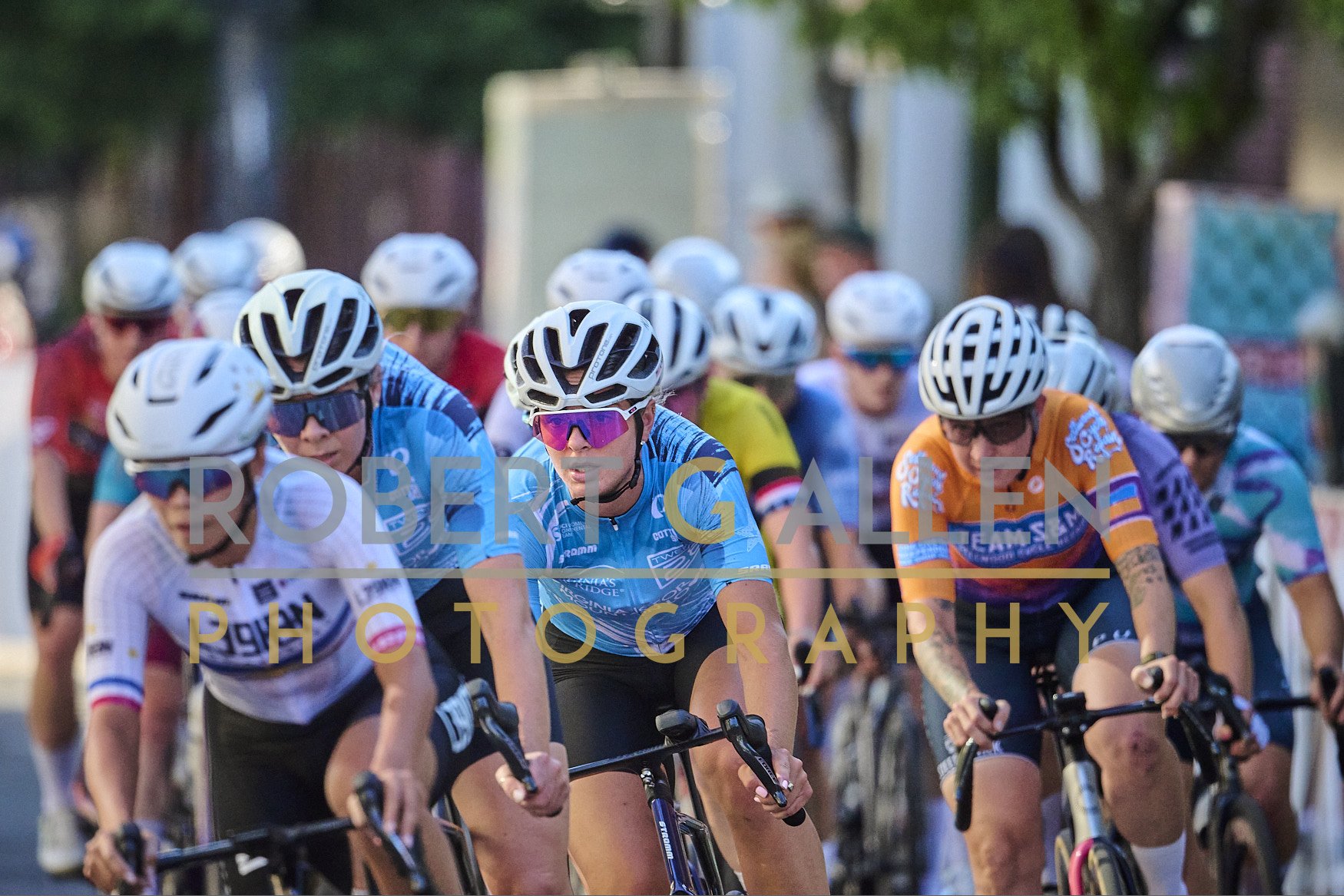 A group of female cyclists wearing helmets and colorful jerseys racing on a road with trees in the background.