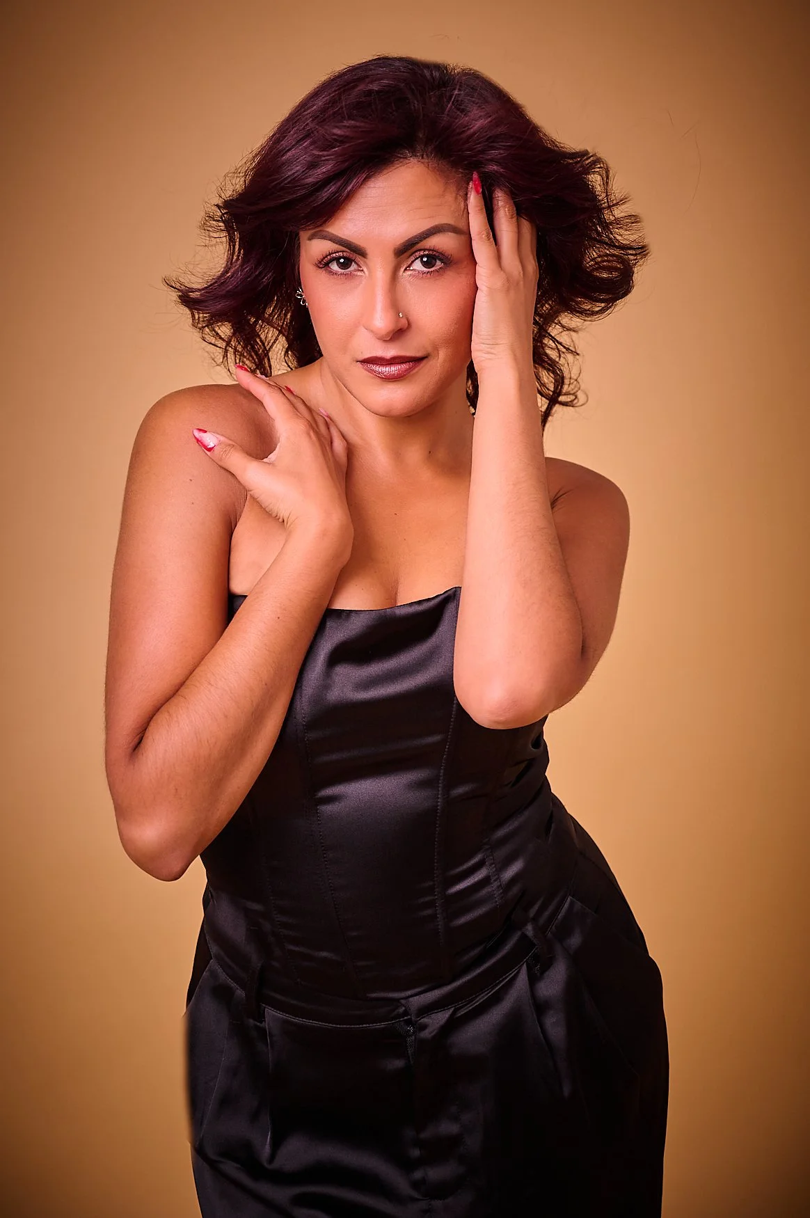 A woman with dark red, wavy hair poses against a plain tan background. She is wearing a strapless black satin dress and has one hand on her head and the other on her shoulder, looking directly at the camera.