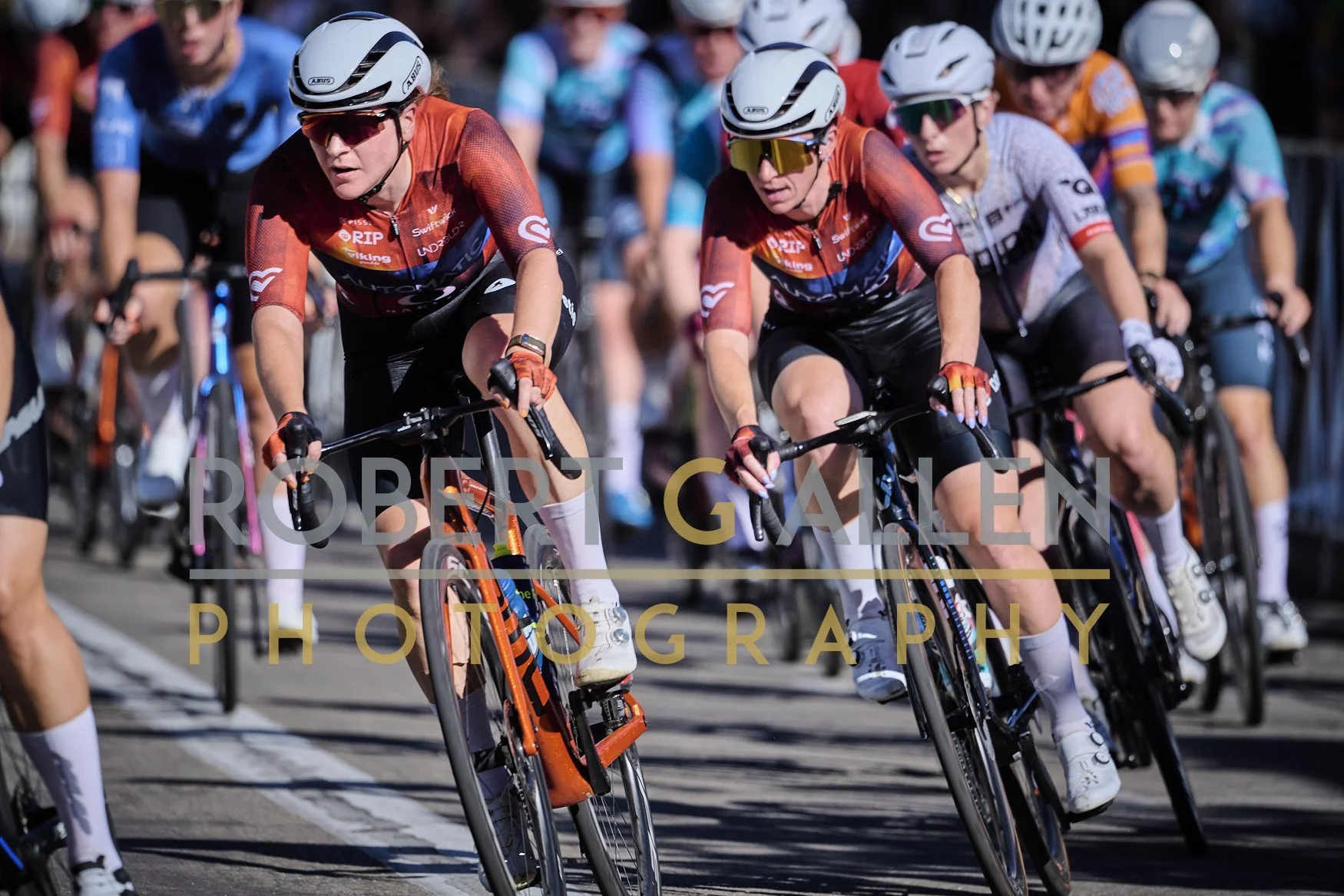 Cyclists participating in a race, riding fast on a city street with focused expressions, wearing helmets and colorful jerseys, with a crowd in the background.