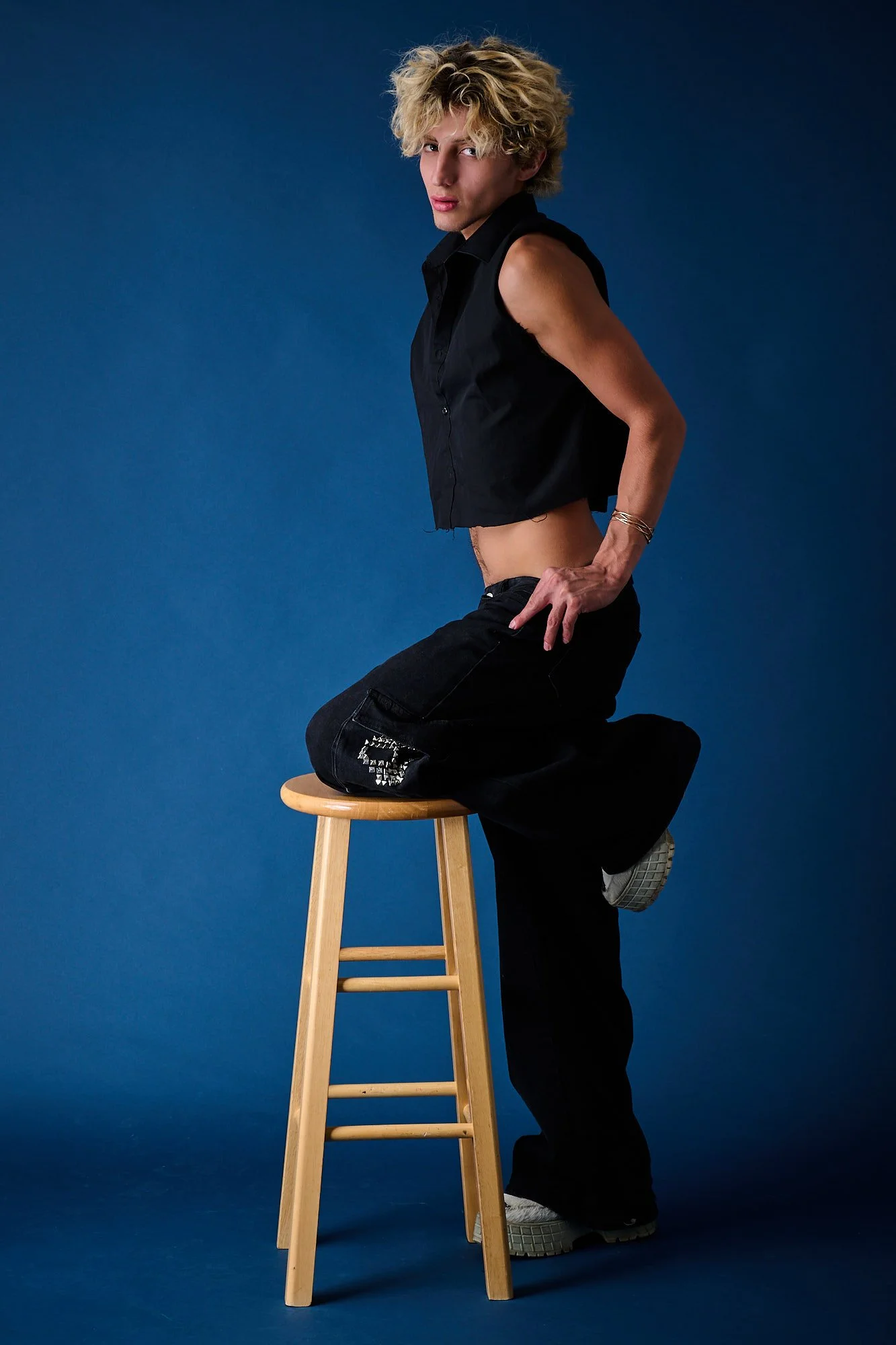 Person with curly blonde hair wearing a black sleeveless shirt and black pants, sitting on a stool with one knee up, against a blue background.