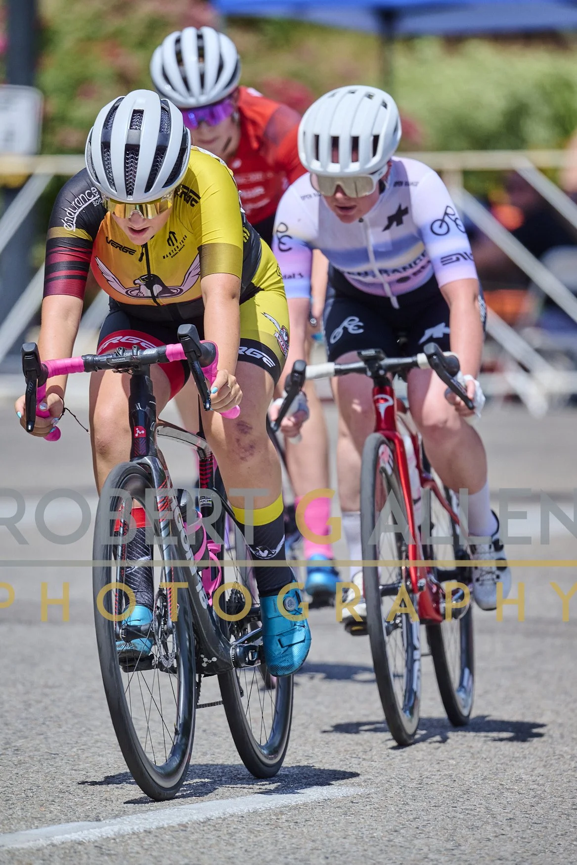 Four female cyclists riding on a road, wearing helmets and sports gear, with the foreground focused on two women in yellow and white jerseys. They seem to be competing in a race.