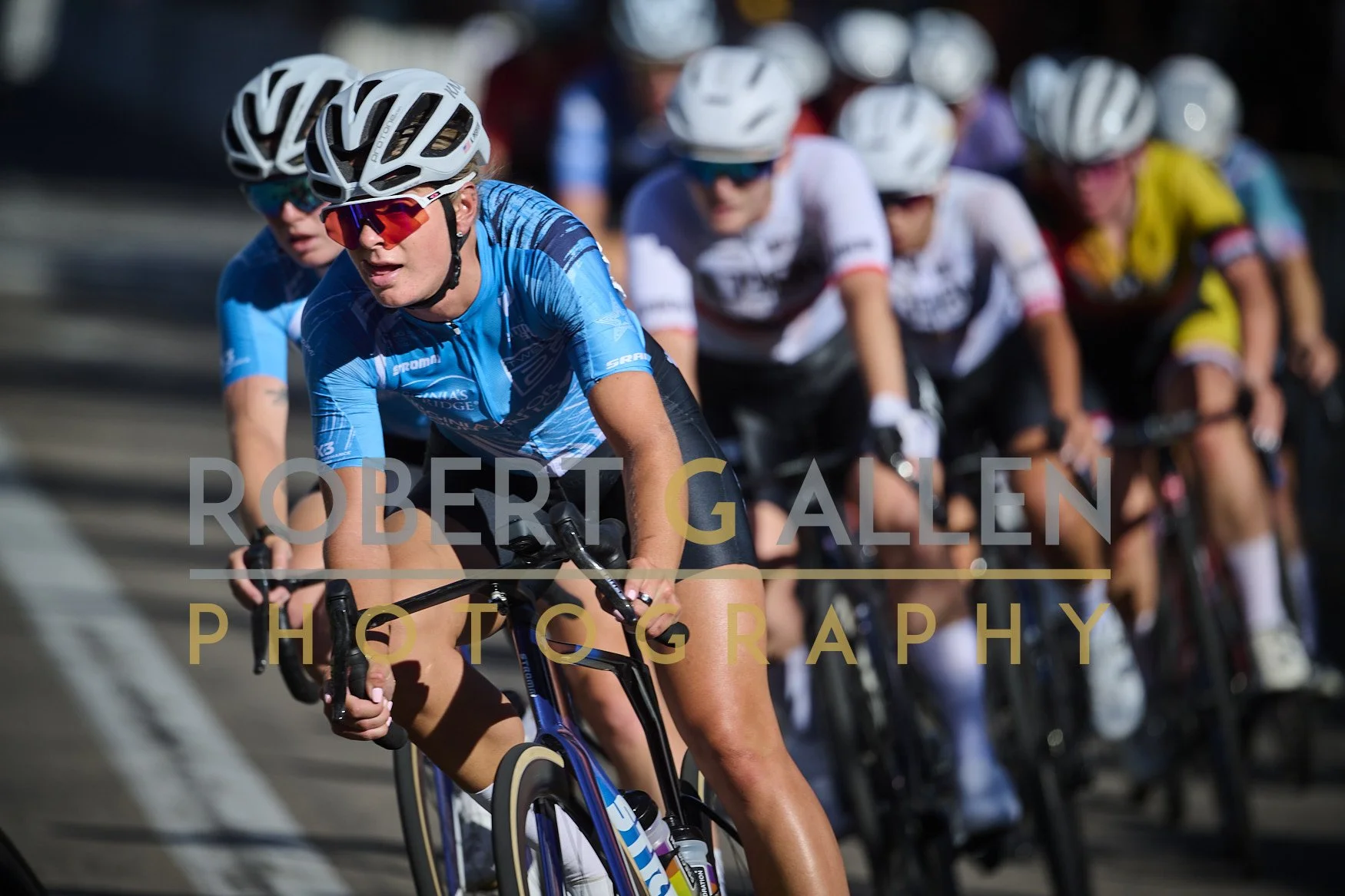Group of women cyclists racing on a road, closely packed, wearing helmets and athletic clothing. The photo is taken from a side angle, showing the intensity of the race.
