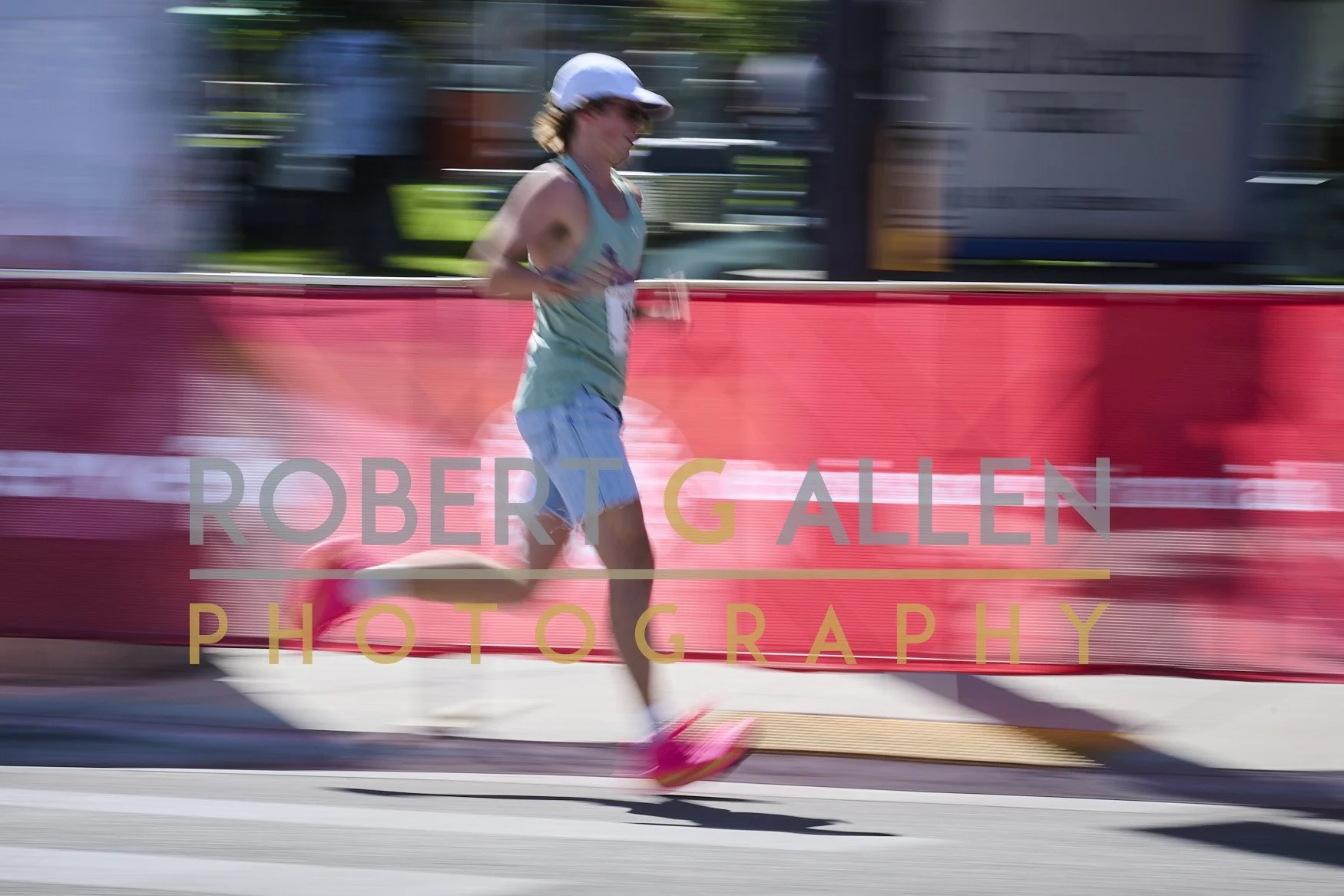 A runner in a blue tank top and shorts, wearing a white visor, participating in a race near a red barrier with the text 'ROBERT G ALLEN PHOTOGRAPHY' overlayed, with a motion-blurred background.