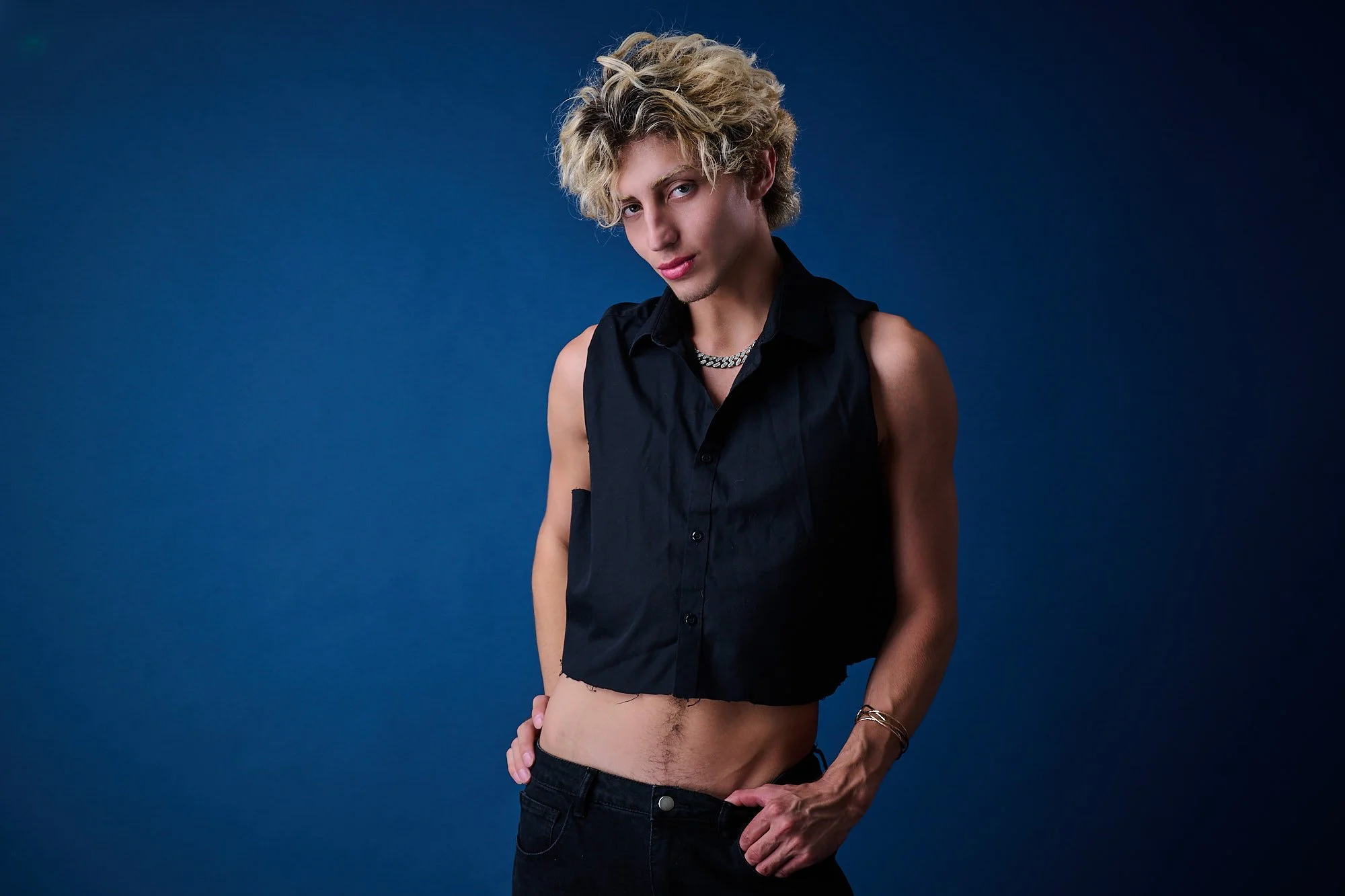 A young man with curly blond hair and light skin wearing a sleeveless black button-up shirt, posing against a dark blue background. His shirt is open, revealing his midriff and some chest jewelry, and he looks confidently at the camera.