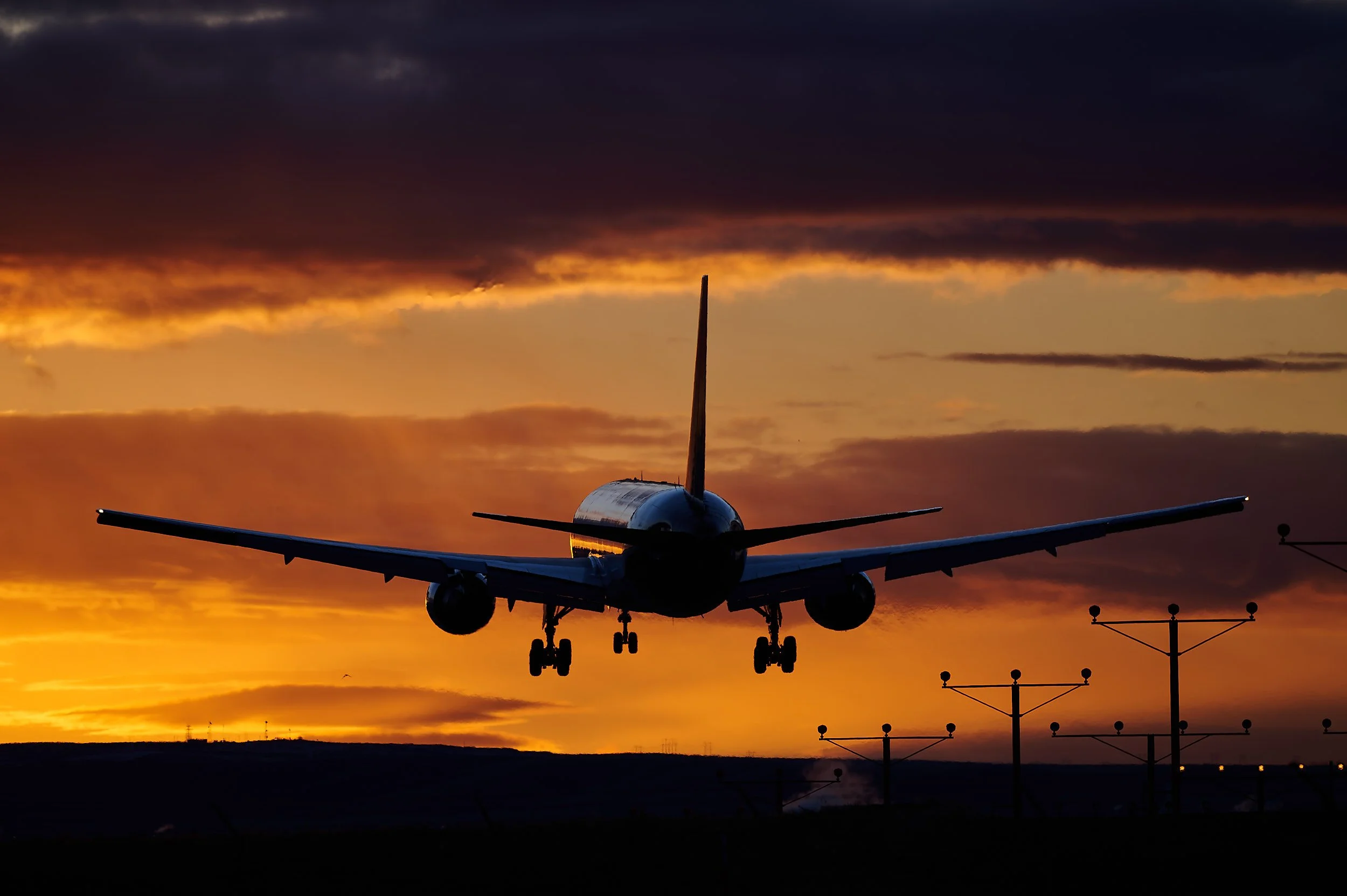 An airplane approaching the runway at sunset with an orange and dark sky and airport lights in the foreground.